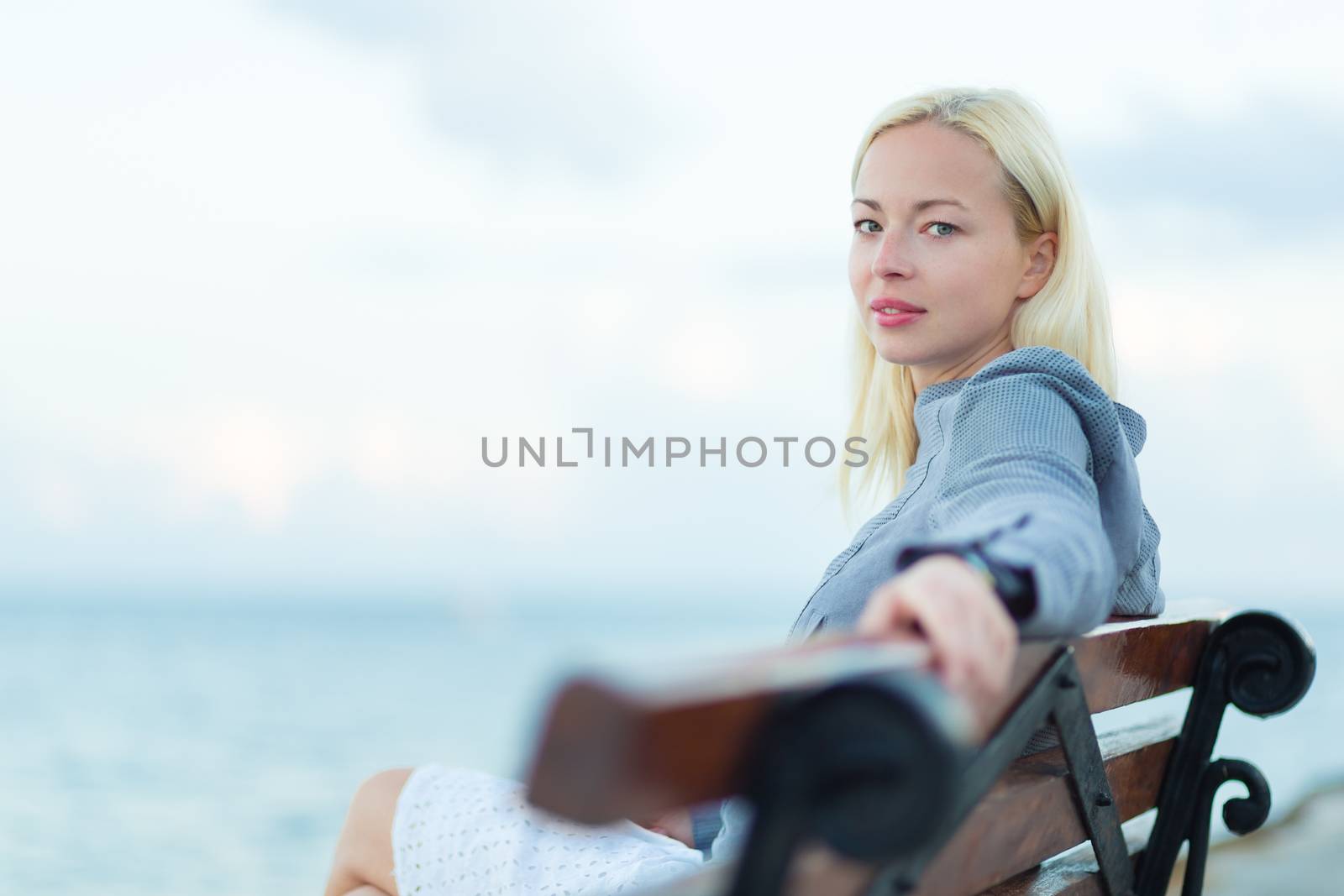 Beautiful woman sitting on a vintage wooden bench, relaxing on fresh breeze by the sea, looking at camera.