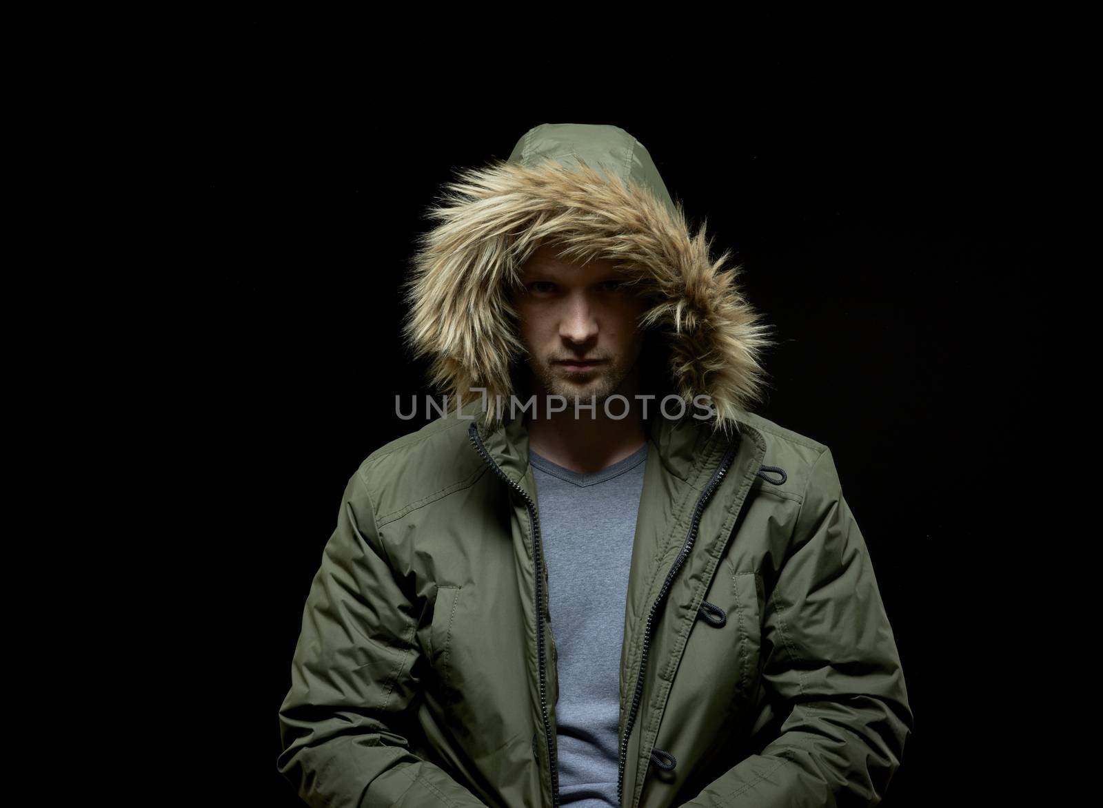 Low key studio portrait of suspicious young adult caucasian model wearing winter coat with hood on. Isolated on black.
