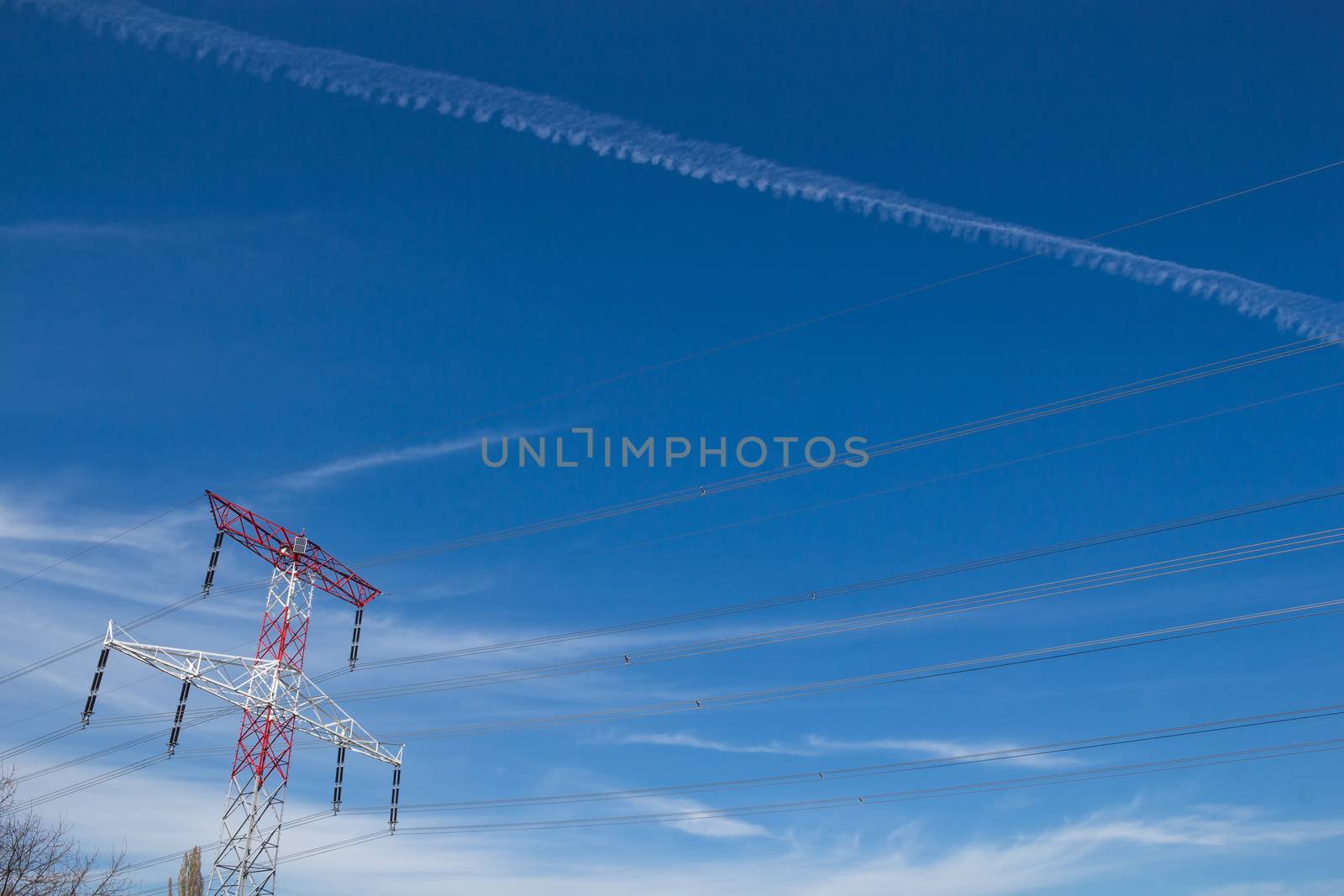 Electricity tower and its cables. Bright blue sky with clouds and a trail.