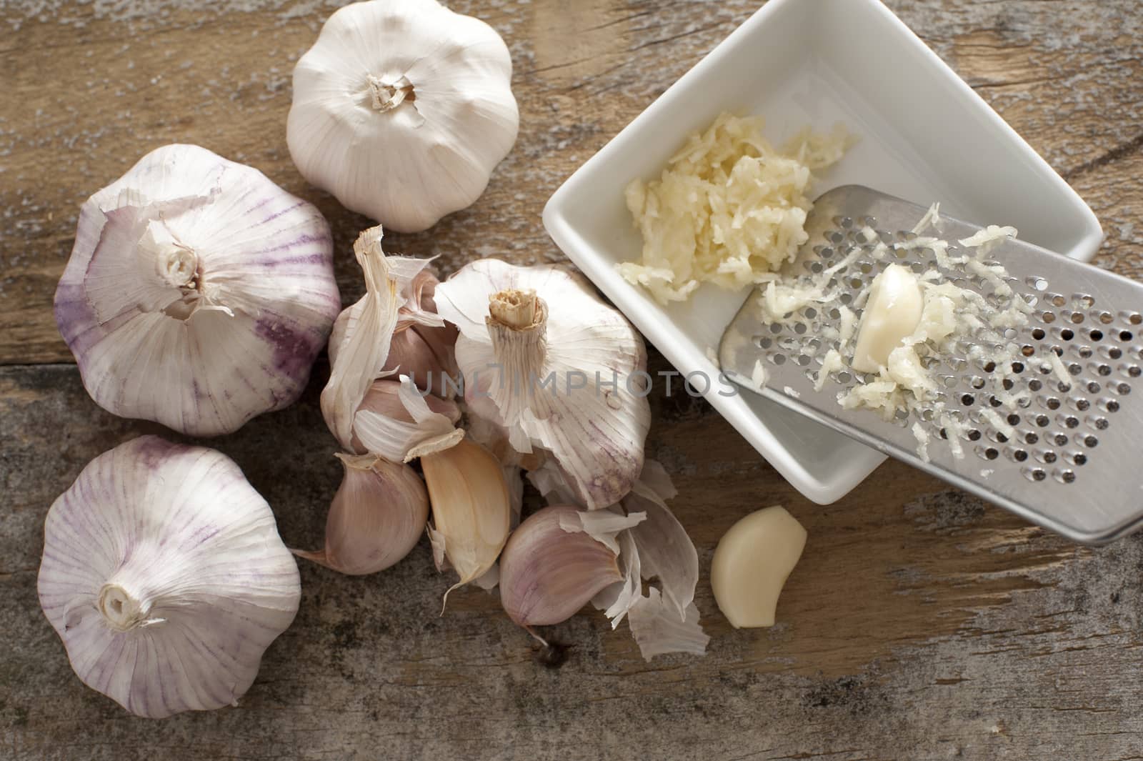 Four garlic bulbs beside a white square bowl holding shredded cloves and stainless steel grater