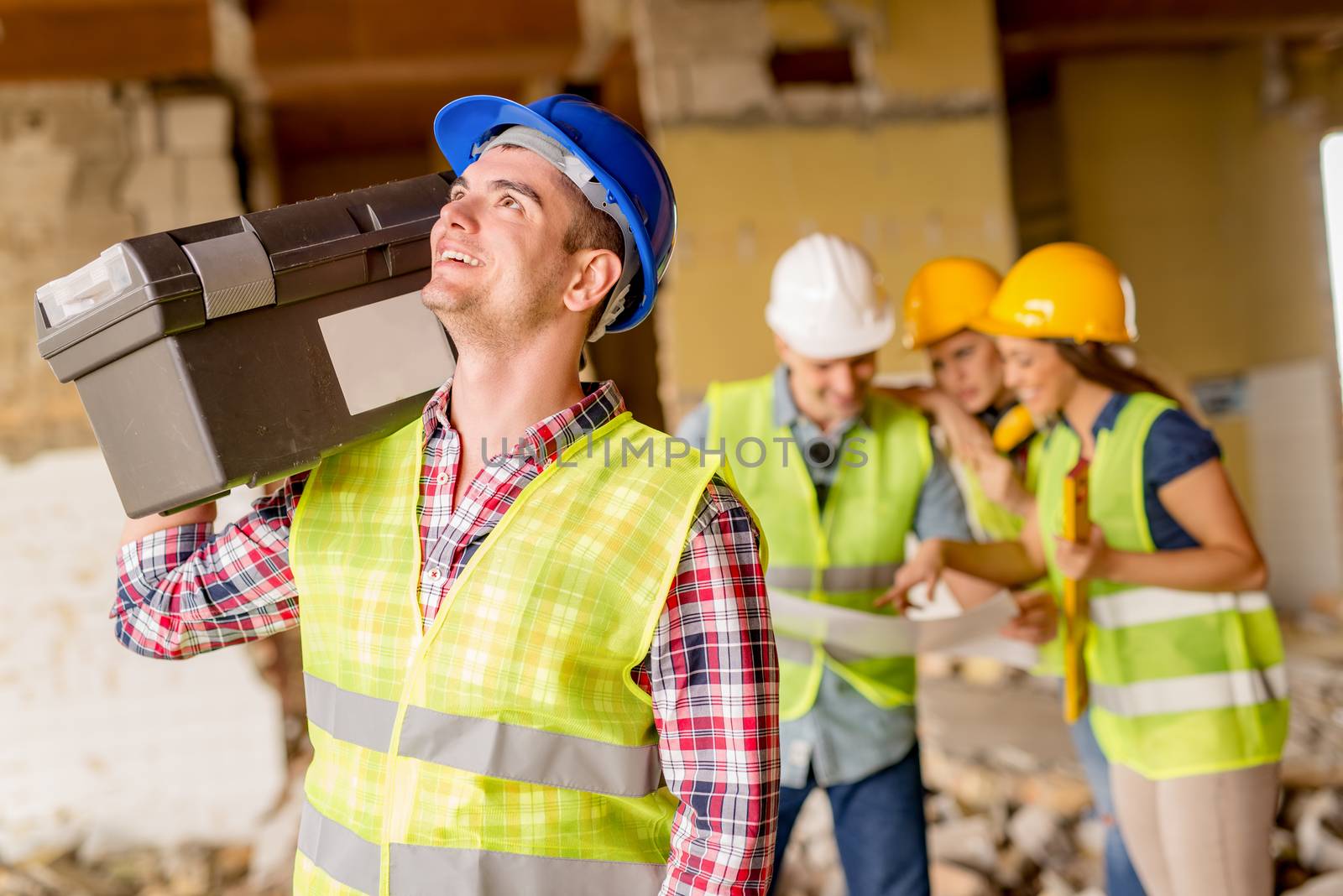 Smiling young electrician with helmet holding toolbox at a construction site. His architect colleagues review plan in background.