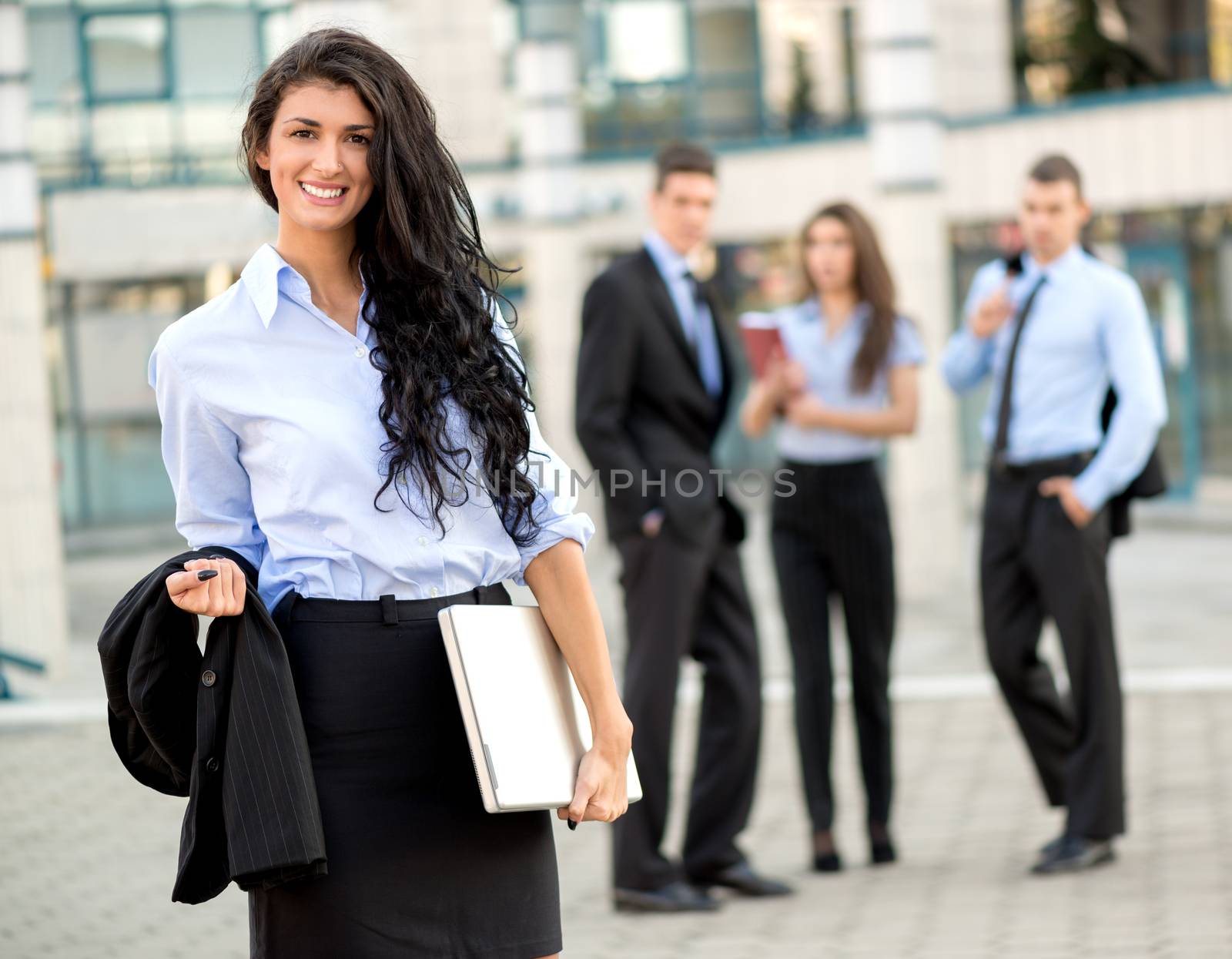 A young businesswoman standing in front of office building separated from the rest of the business team, holding laptop and looking at the camera.