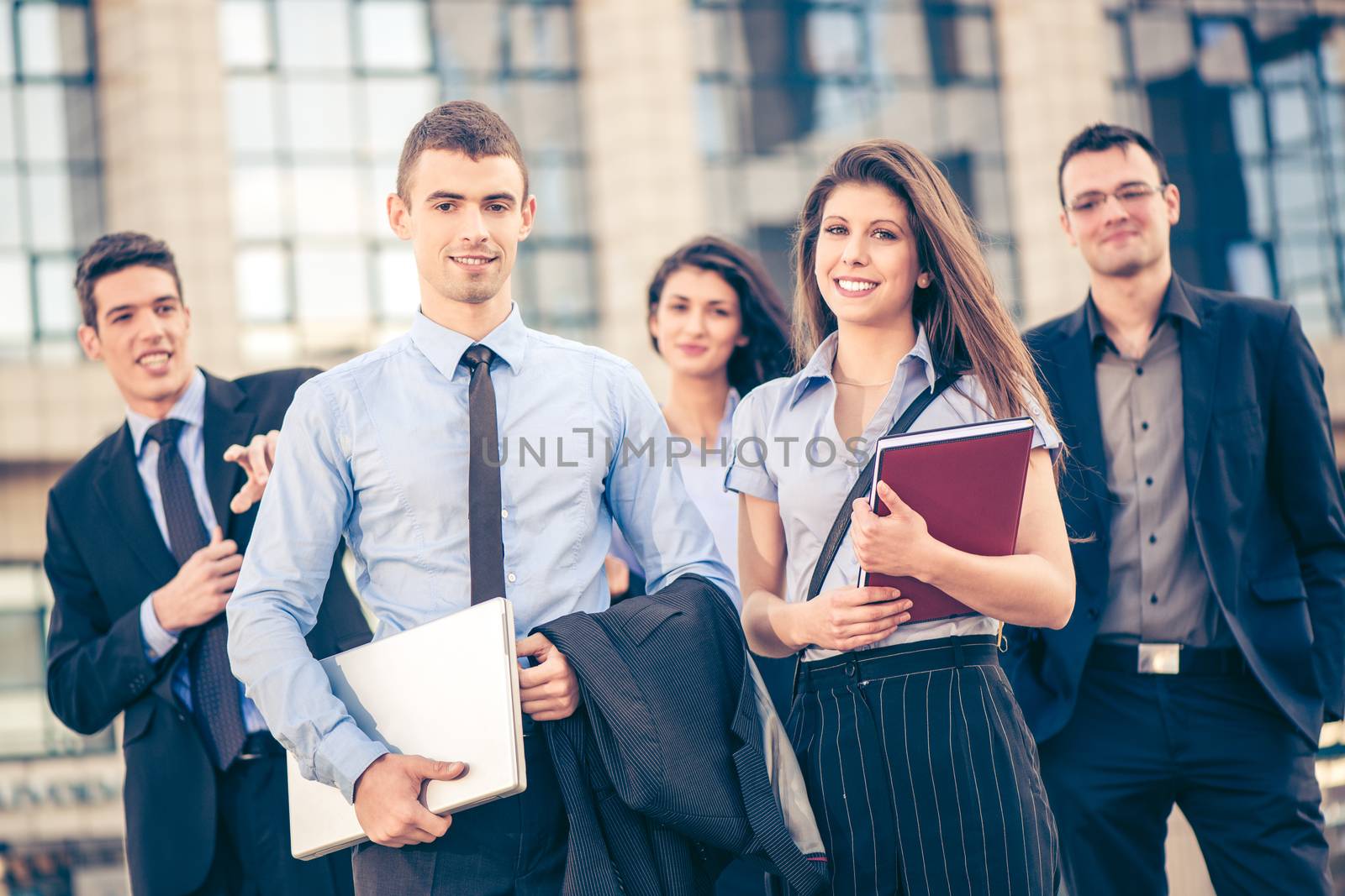 Portrait of group of young successful business people dressed in suits, standing in front of office building, smiling and looking at camera.