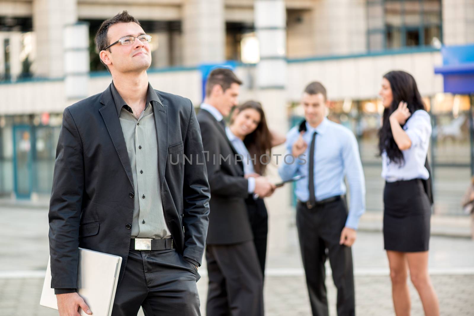 Young businessman with a laptop standing in front of office building separated from the rest of the business team. With a smile looking into the distance.