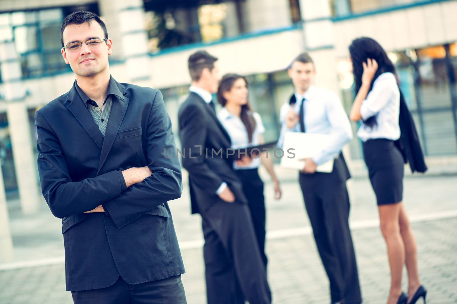 Young businessman standing in front of office building separated from the rest of the business team. With a smile looking at the camera.