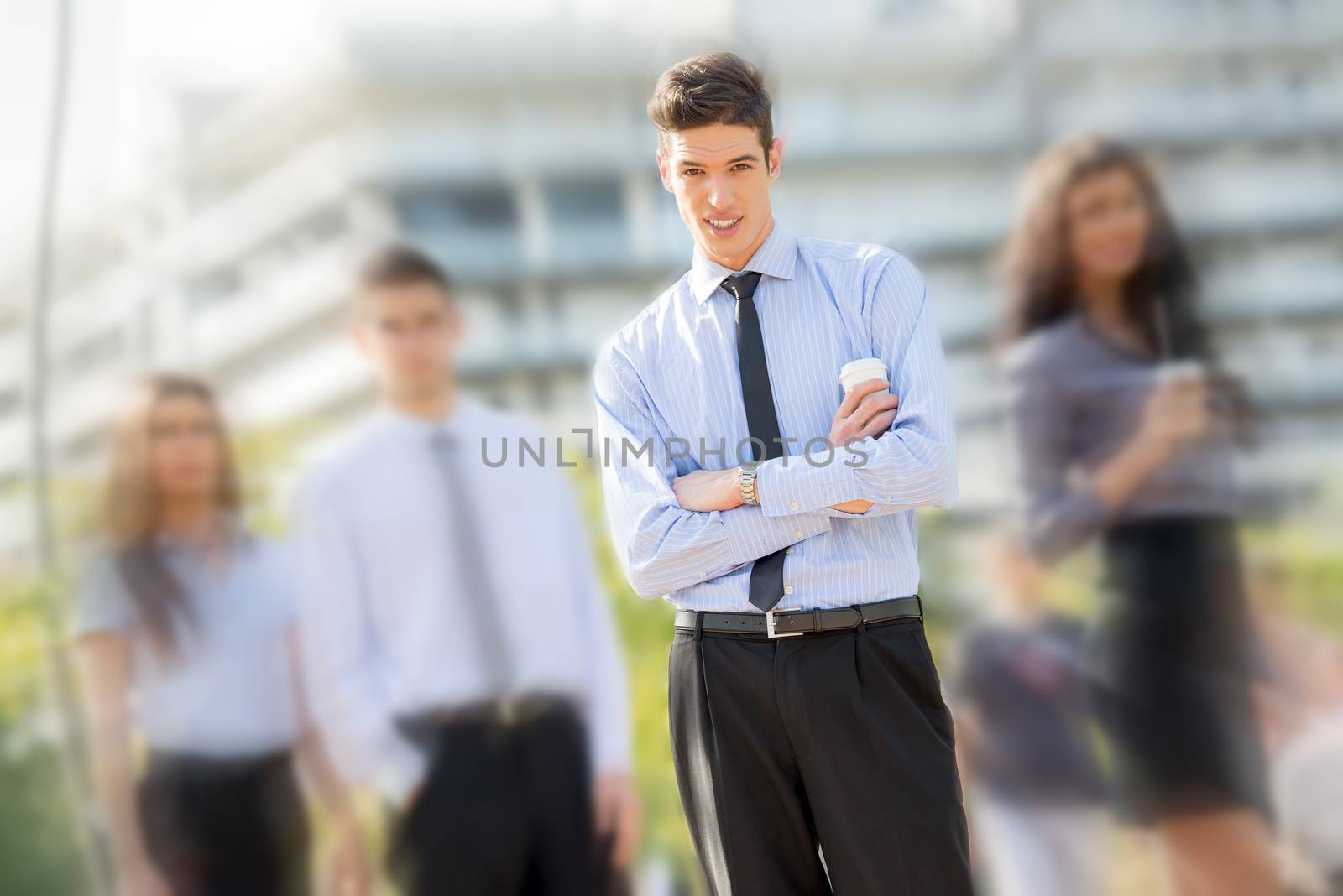 Young businessman, elegantly dressed, standing  with his team on a coffee break in front of office building and looking at the camera with.