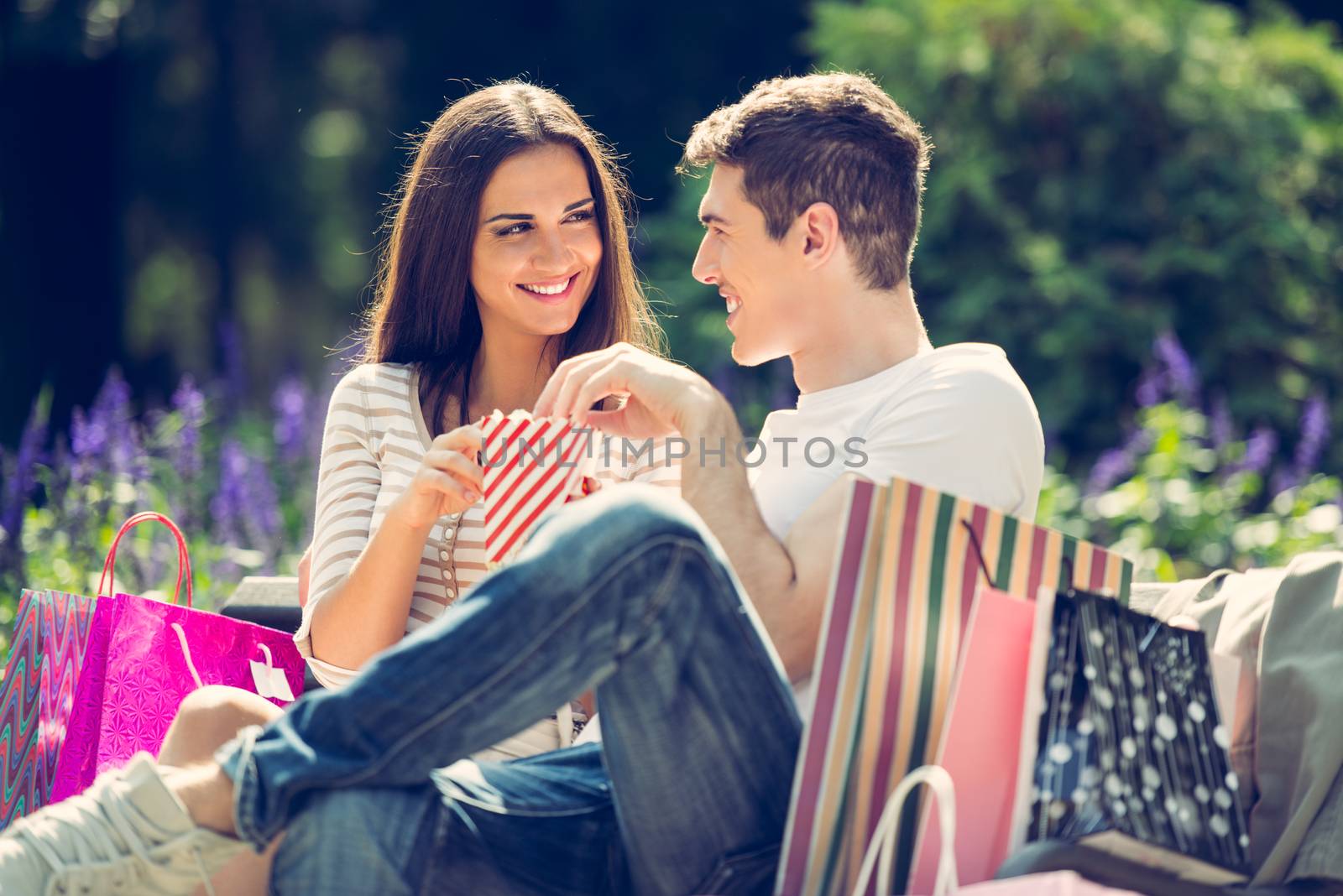 A young loving couple resting on a park bench after shopping, enjoying the sun while nibbling popcorn.