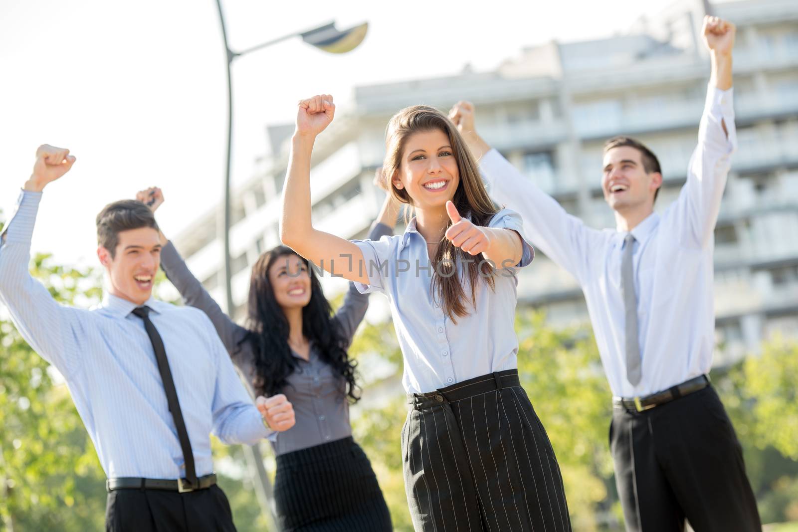 Young businesswoman elegantly dressed with his team celebrating business success at a park near his office building, arms raised and outstretched thumb on looking at the camera.