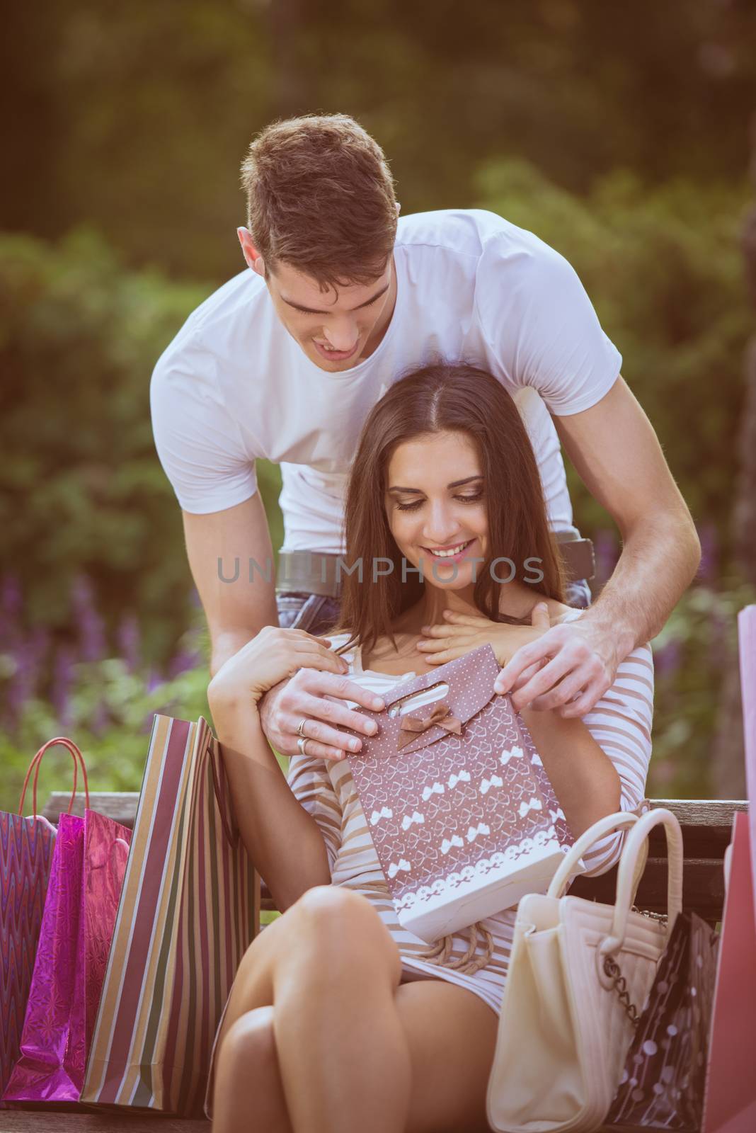 A young girl sitting on a park bench enjoying the beautiful sunny day, resting from shopping in addition to a bunch of shopping bags, her boyfriend holding a gift bag  and behind the bench leaning toward her face.