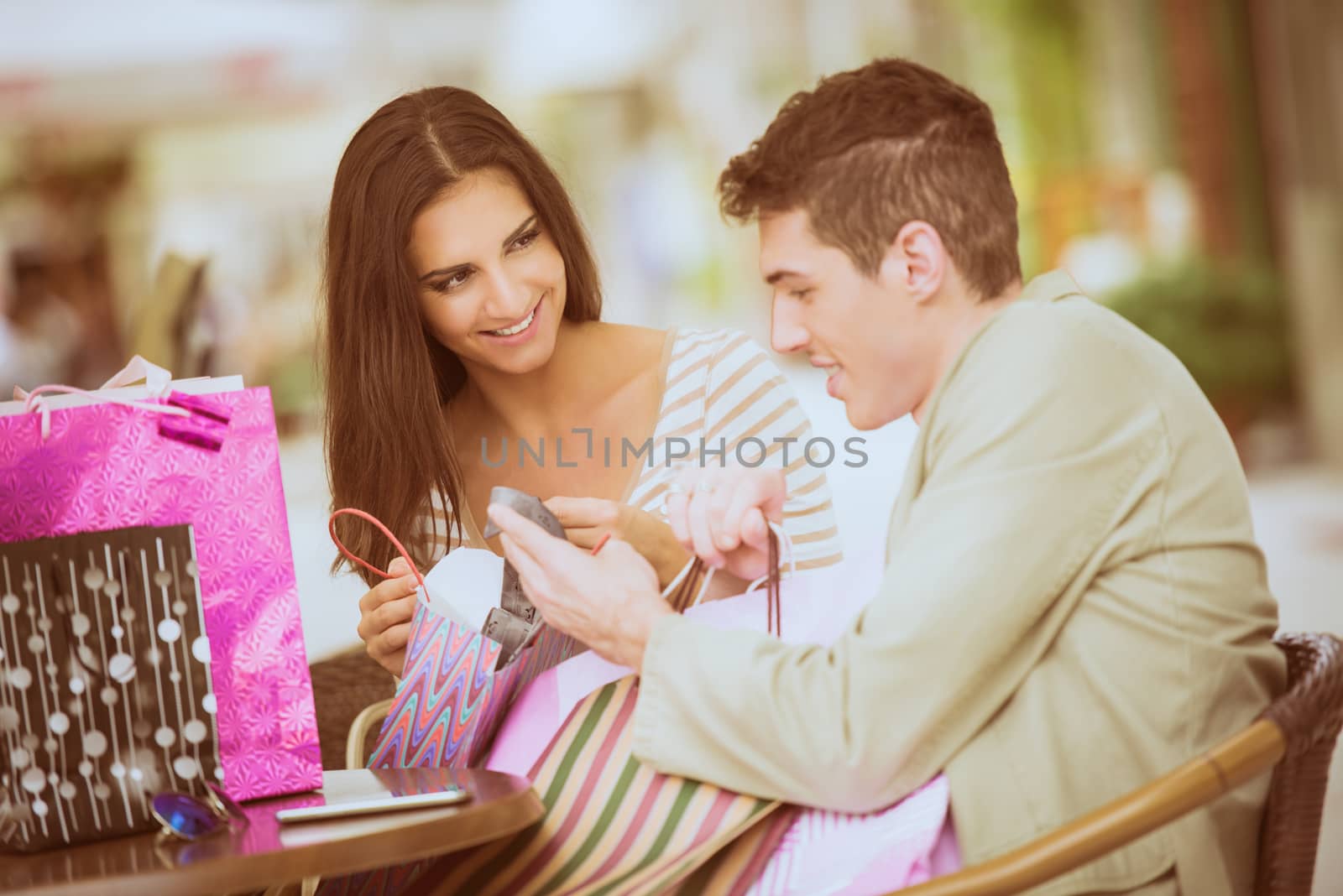 Girl and her boyfriend are resting after shopping in outdoor cafe with colorful chairs, watching with a smile in shopping bags, excited about the things they bought.