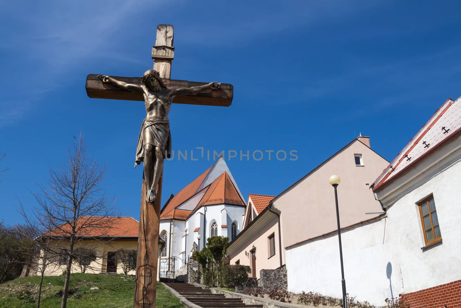 Traditional slovak village, built on a hill, with a church on the top of it. Wooden cross beside the stairs to the gothic church. Springtime with a green grass and blue sky.