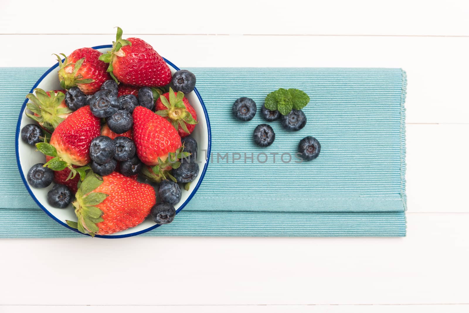 Ceramic bowl with blueberries and strawberries at blue textile napkin over wooden table. Top view with copy space