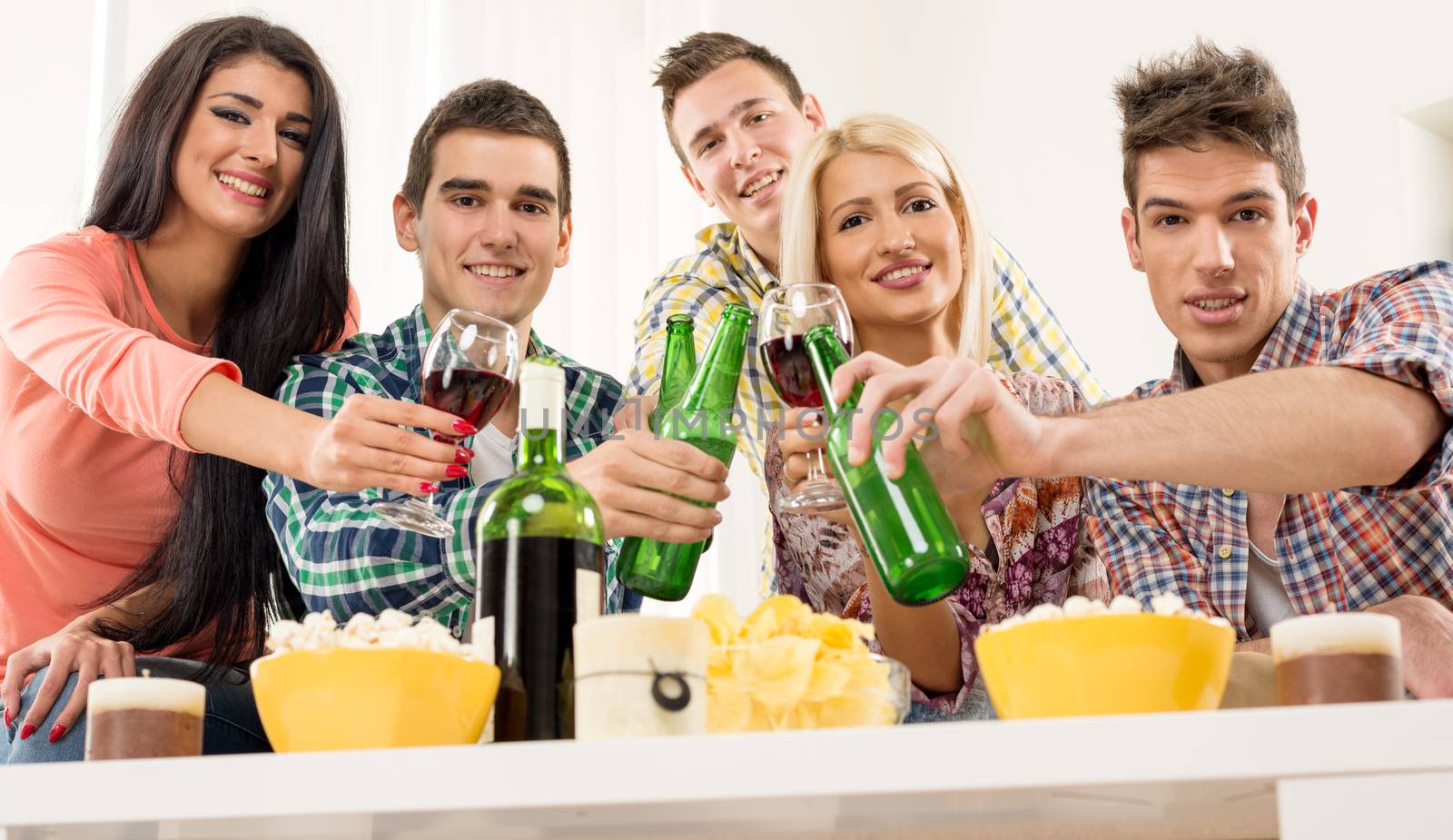 Cheerful friends at home party toasting knocking with bottles and glasses. With a smile looking at the camera.