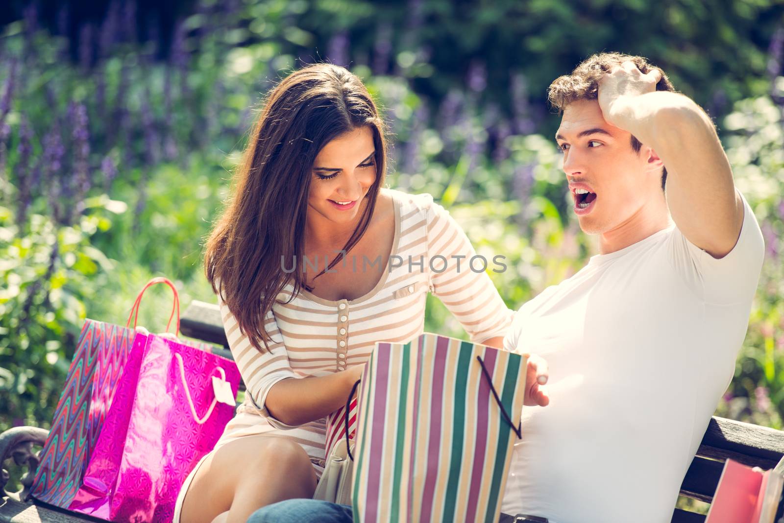 Young guy with a worried expression on his face and his smiling girlfriend who sits on a park bench next to a bunch of shopping bags.