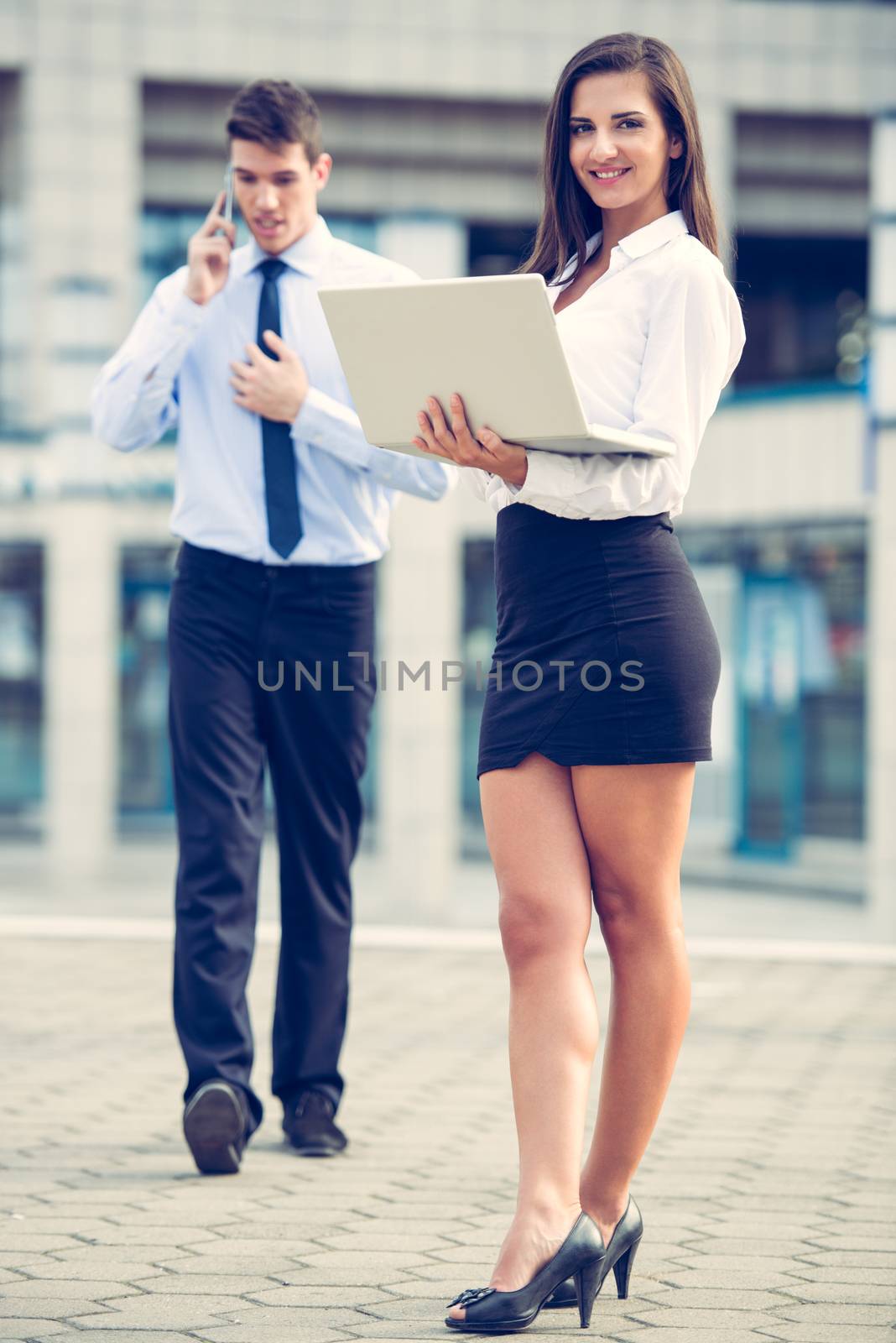 Young business partners standing in front of the building. Business woman working on a laptop while her partner use a mobile phone for business conversation.