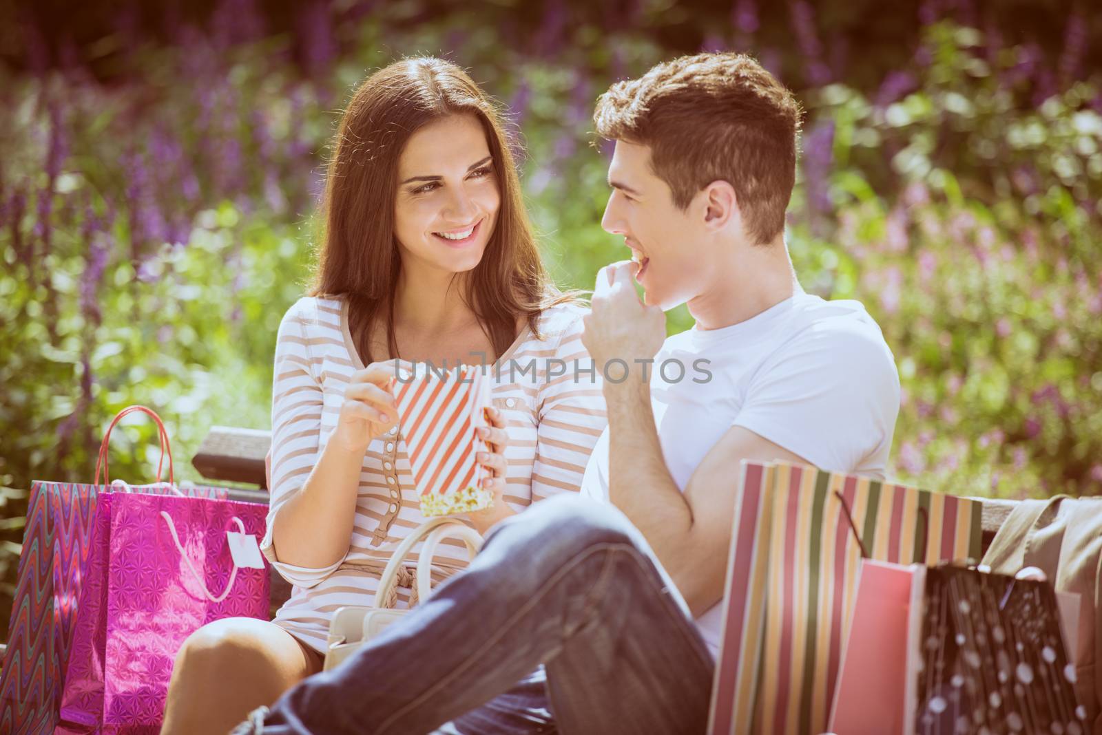 A young loving couple resting on a park bench after shopping, enjoying the sun while nibbling popcorn.