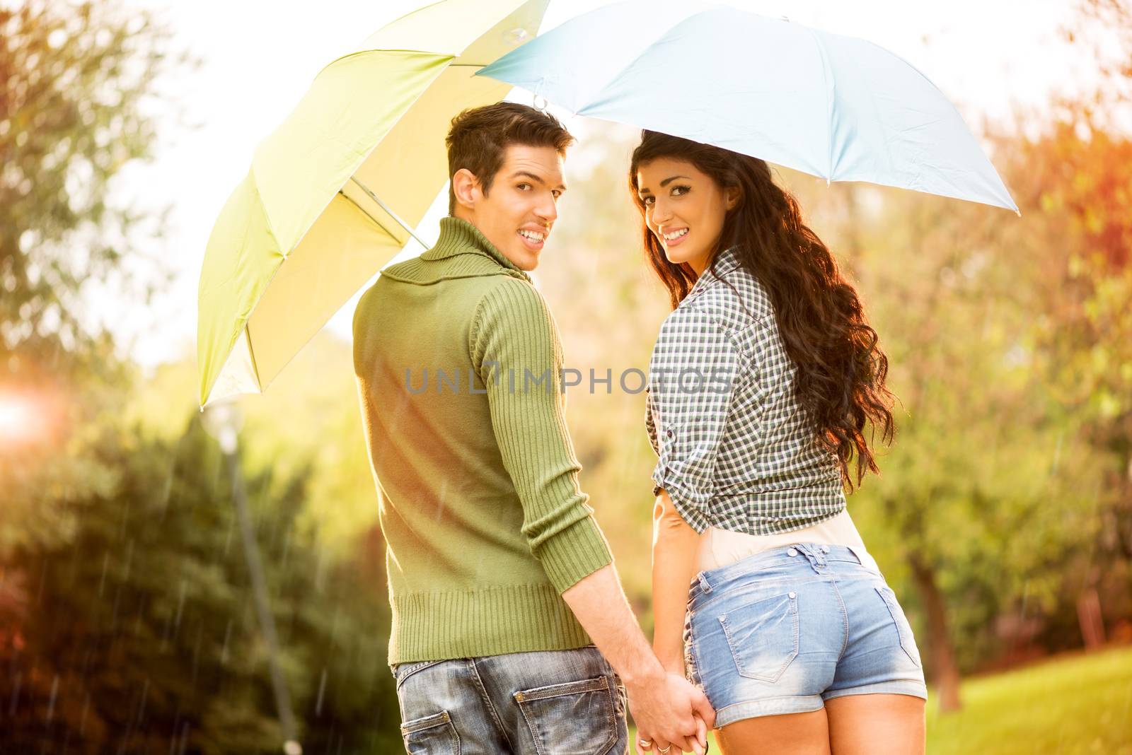 Rear view of a young couple in love with umbrellas while walking in the rain through the park holding hands and looking at camera other with a smile.