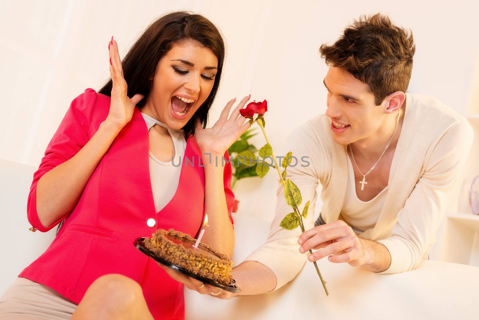 Young man with a rose in his mouth gives beautiful brunette girl birthday cake, pleasantly surprised girl with wide open mouth looking into the cake.