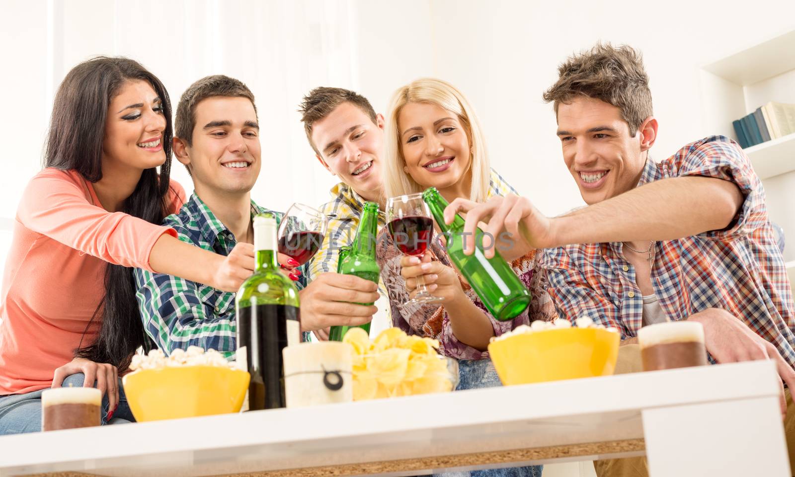 Friends at home party smiling and cheerful toast knocking with bottles and glasses.