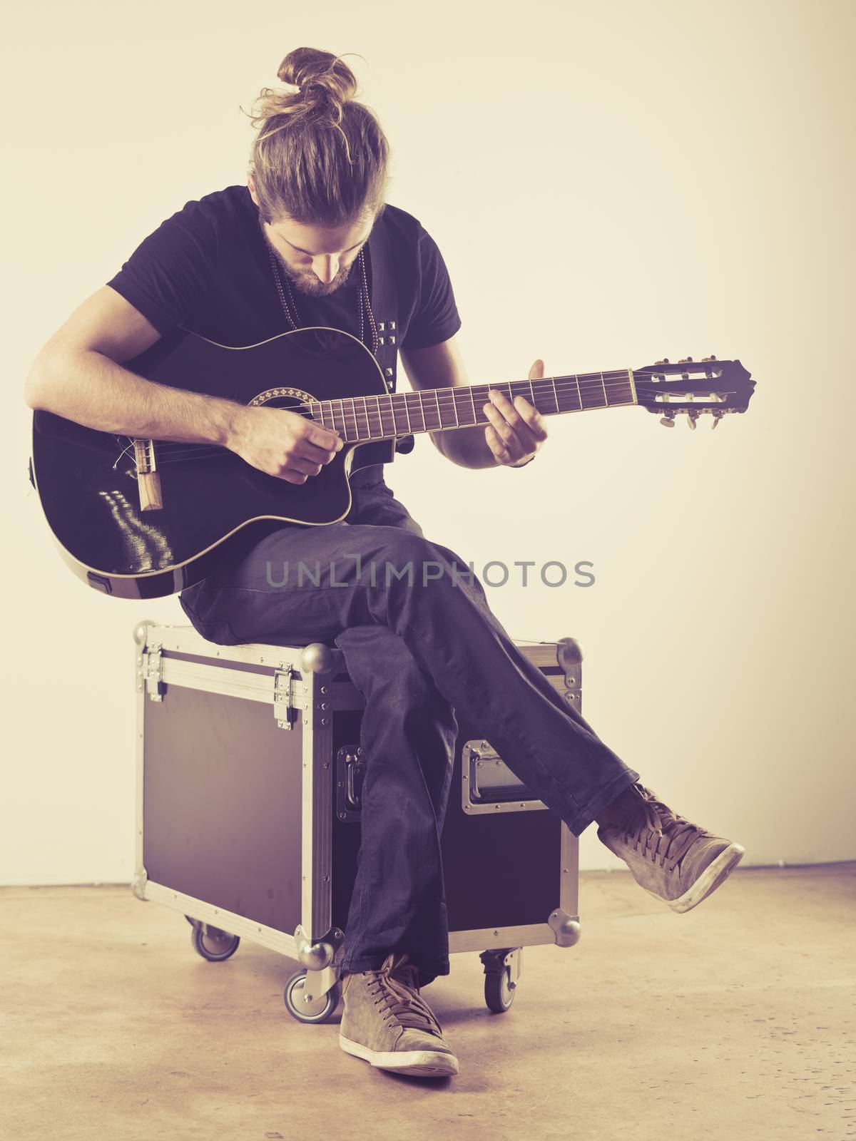Photo of a young attractive man with long hair and beard sitting on a flight case and playing an acoustic guitar. Filtered to look vintage.
