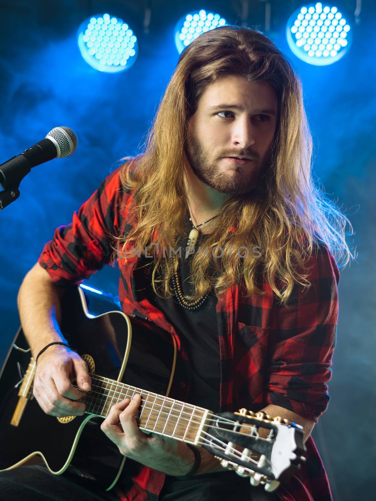 Photo of a young man with long hair and a beard playing an acoustic guitar on stage with lights and concert atmosphere.
