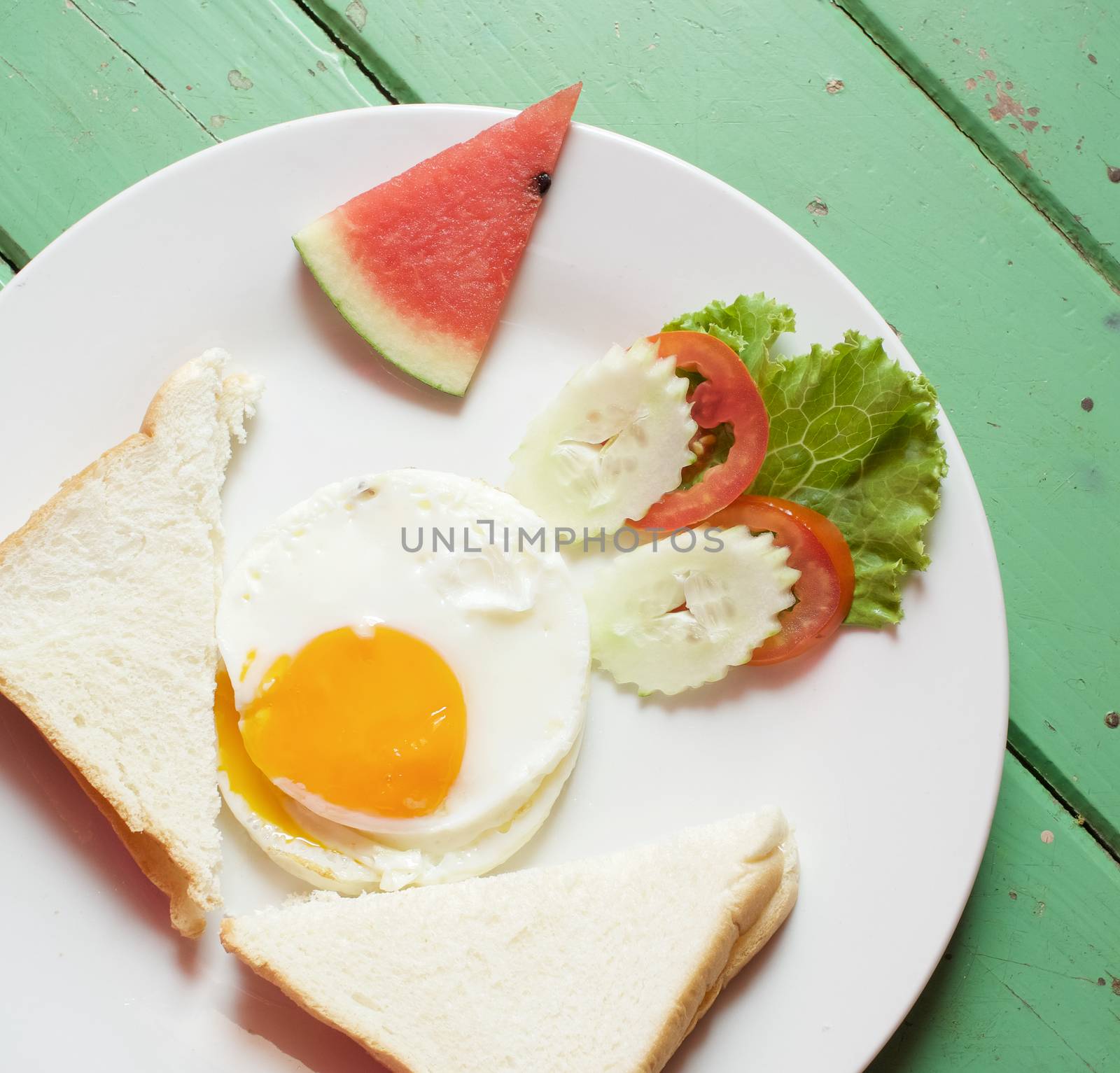 Fried egg, bread, cucumber, tomato and lettuce with watermelon in white plate on green wood table.