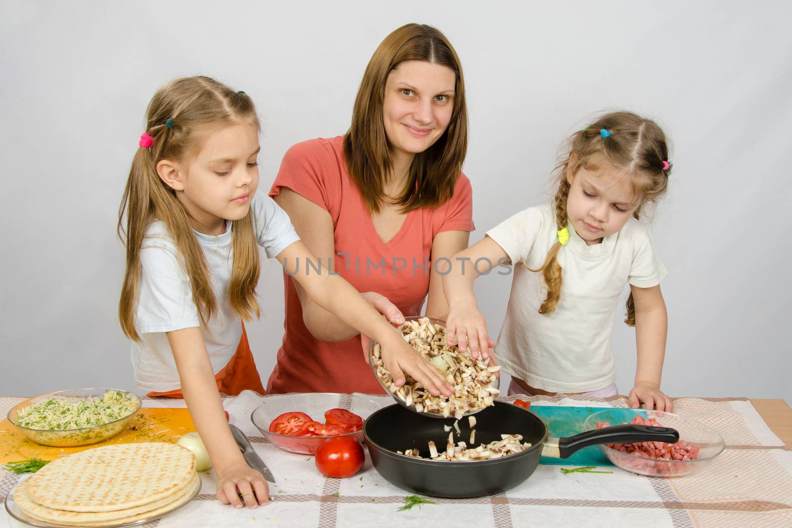 Two little girls at the kitchen table with a zeal to help her mother pour the mushrooms from the plate to the pan