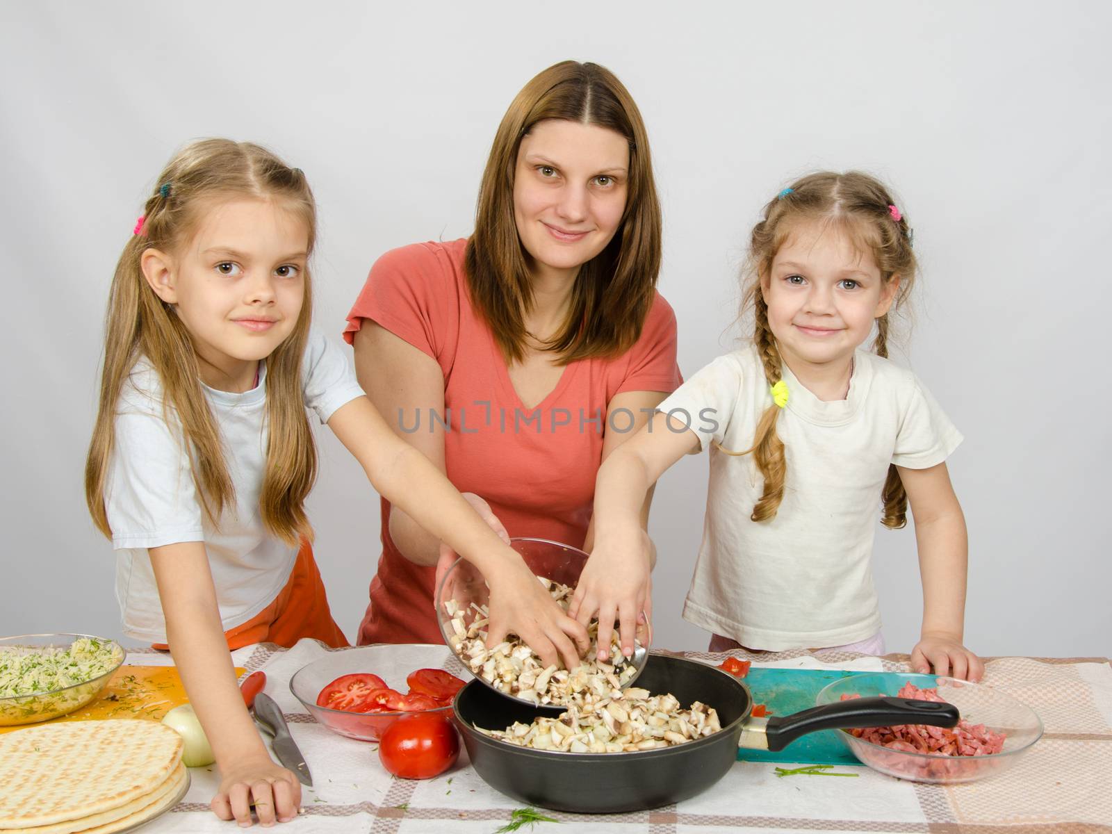 Two little daughter at the kitchen table helping her mother pour chopped mushrooms from the plate to the pan