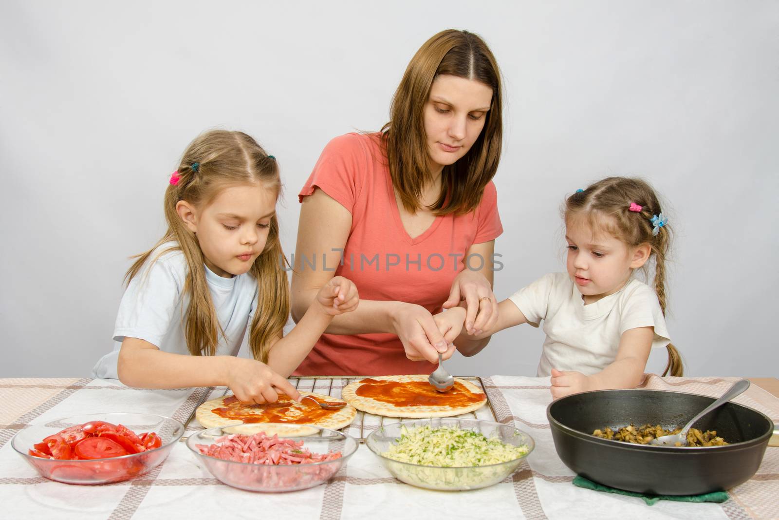 Mom helps younger daughter spread ketchup on a pizza, the eldest daughter, she is preparing a second pizza