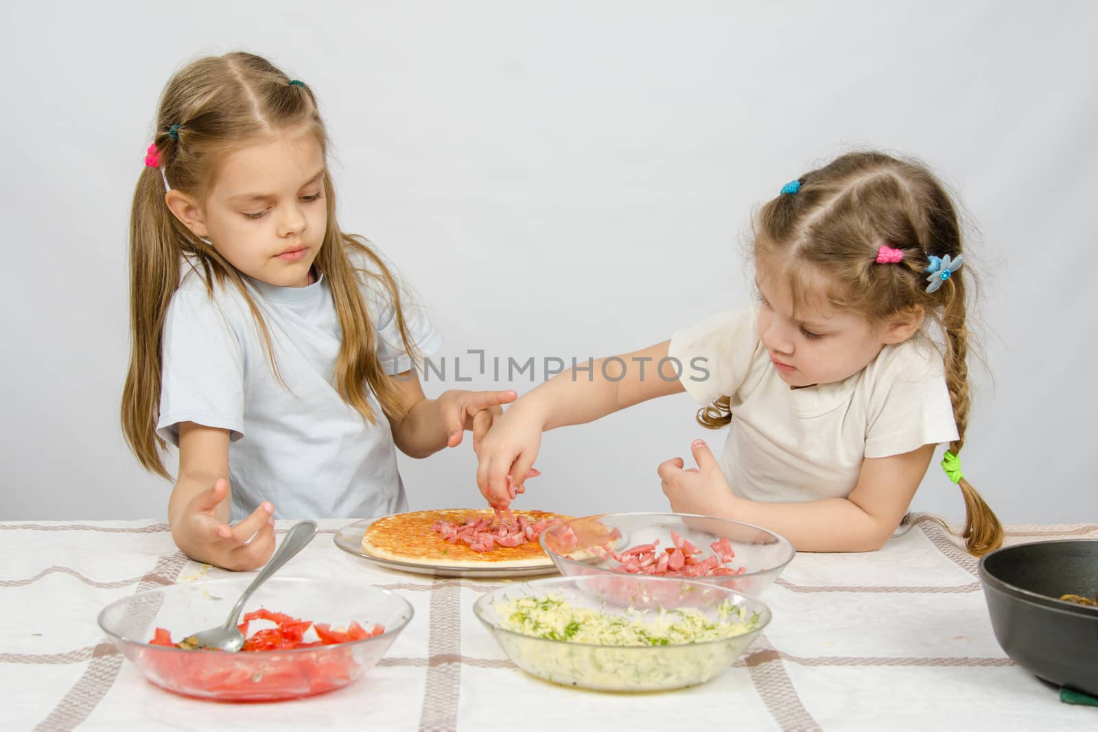 Six-year girl observes and controls her younger sister puts on the pizza ingredients