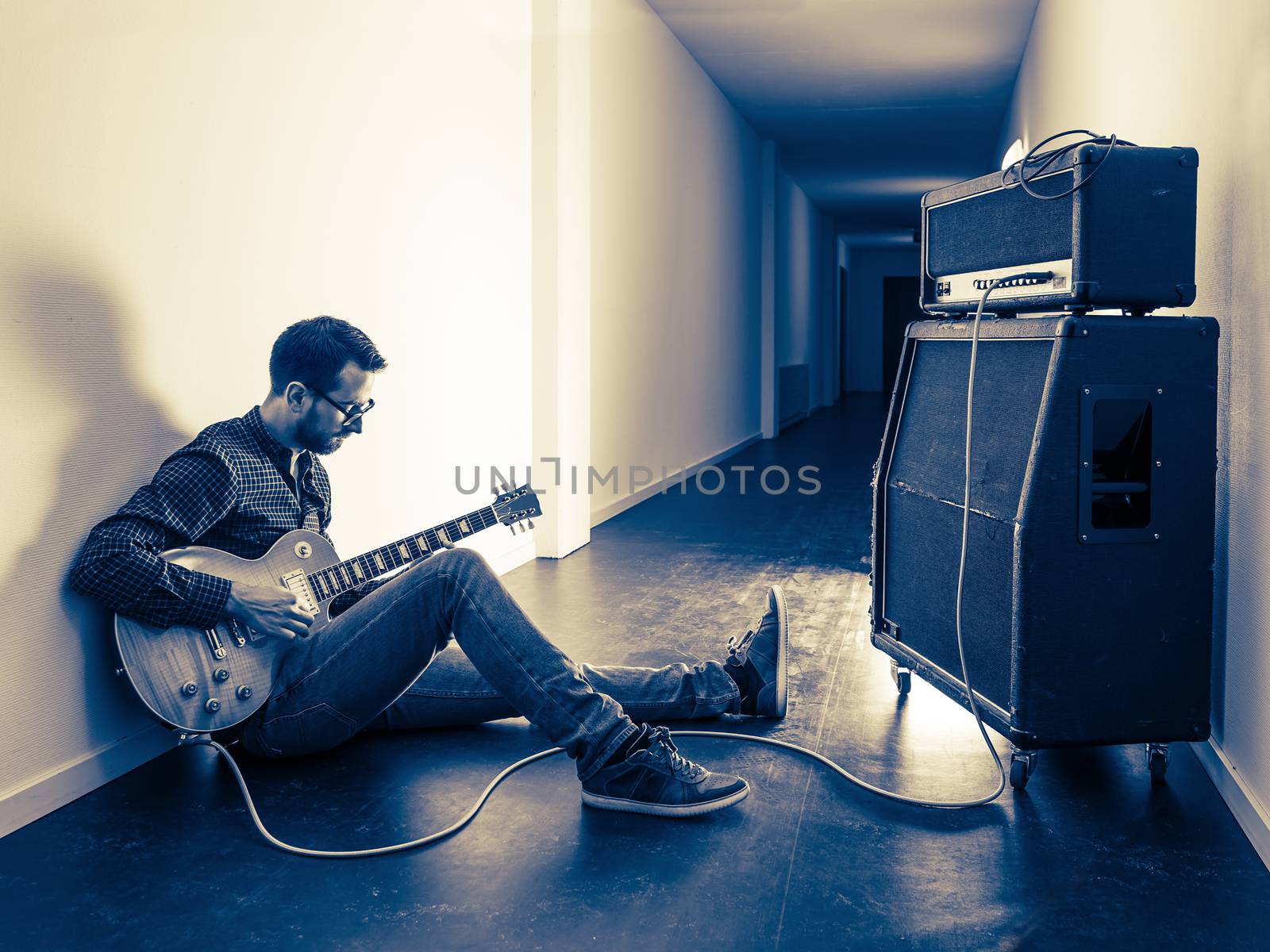 Photo of a man sitting playing his electric guitar in front of a large amplifier in a hallway. Filtered for retro vintage feel.
