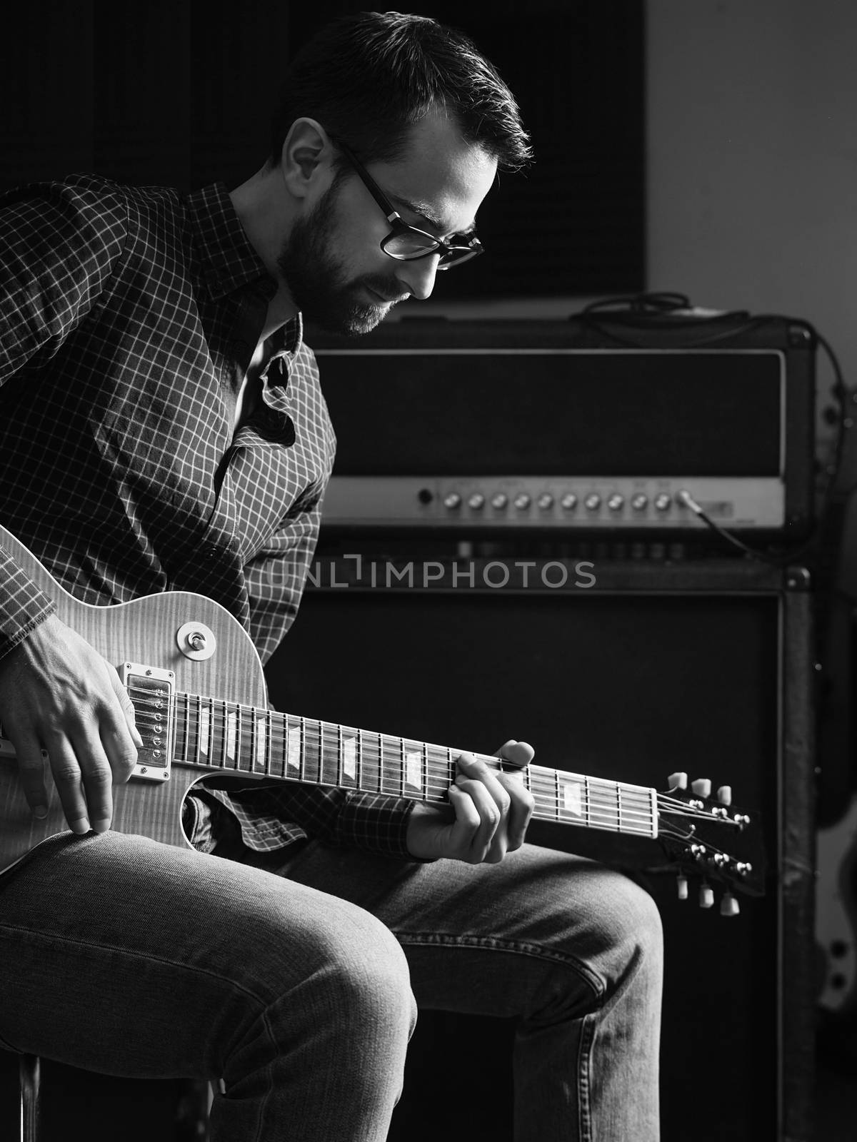 Photo of a man sitting playing his electric guitar in front of a large amplifier.
