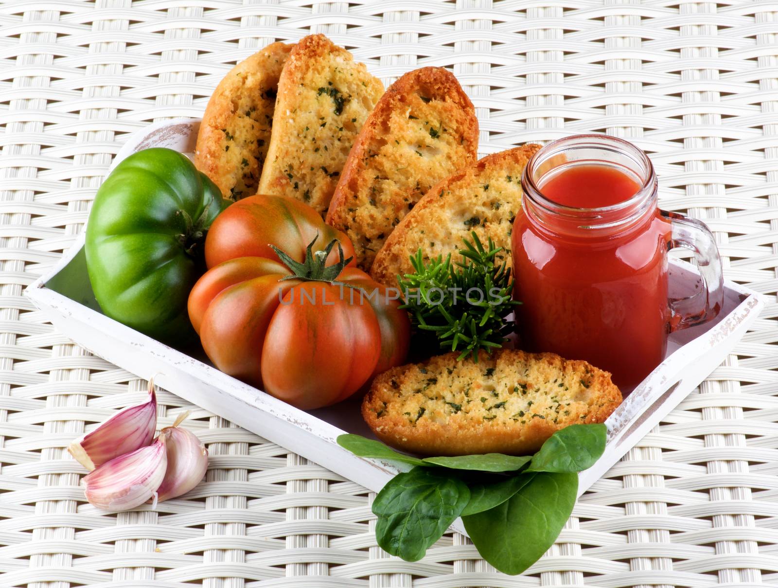Arrangement of Tomato Juice in Glass Jar with Raw Tomatoes, Crispy Bread and Fresh Herbs into White Wooden Tray closeup on Wicker background
