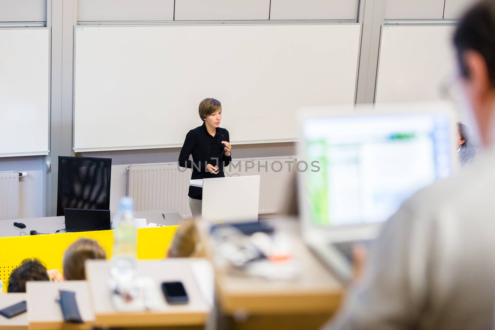 Speaker giving presentation in lecture hall at university. Participants listening to lecture and making notes.