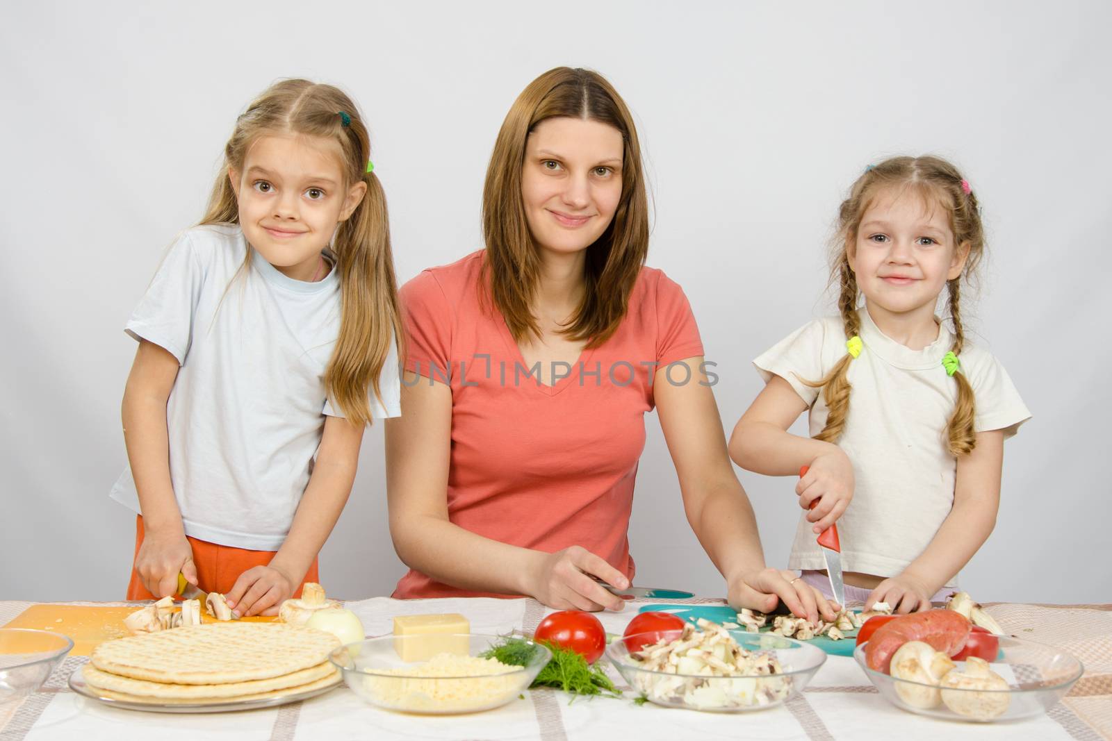 Woman with two young girls at the table prepared ingredients for pizza