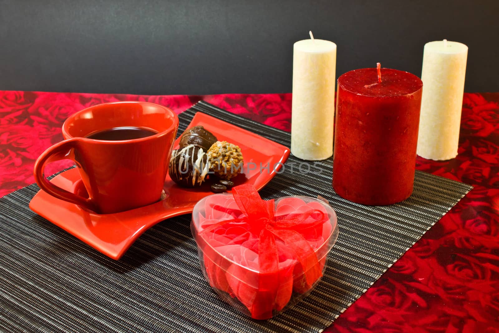 Coffee in red cup on a saucer with a chocolate, a present in a box in the form of heart on the background of candles