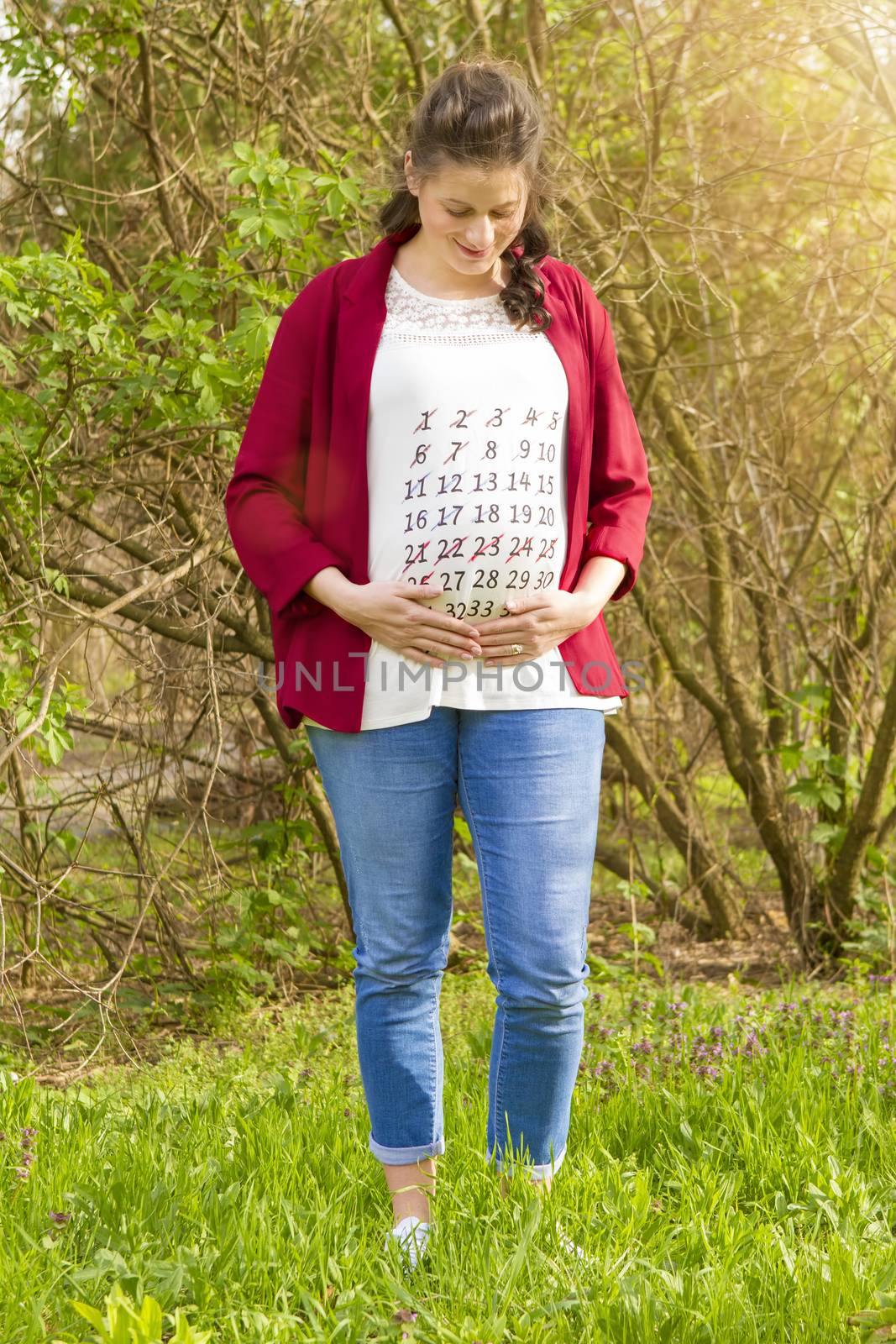Pregnant woman in red jacket with calendar on her T-shirt outdoor in the park
