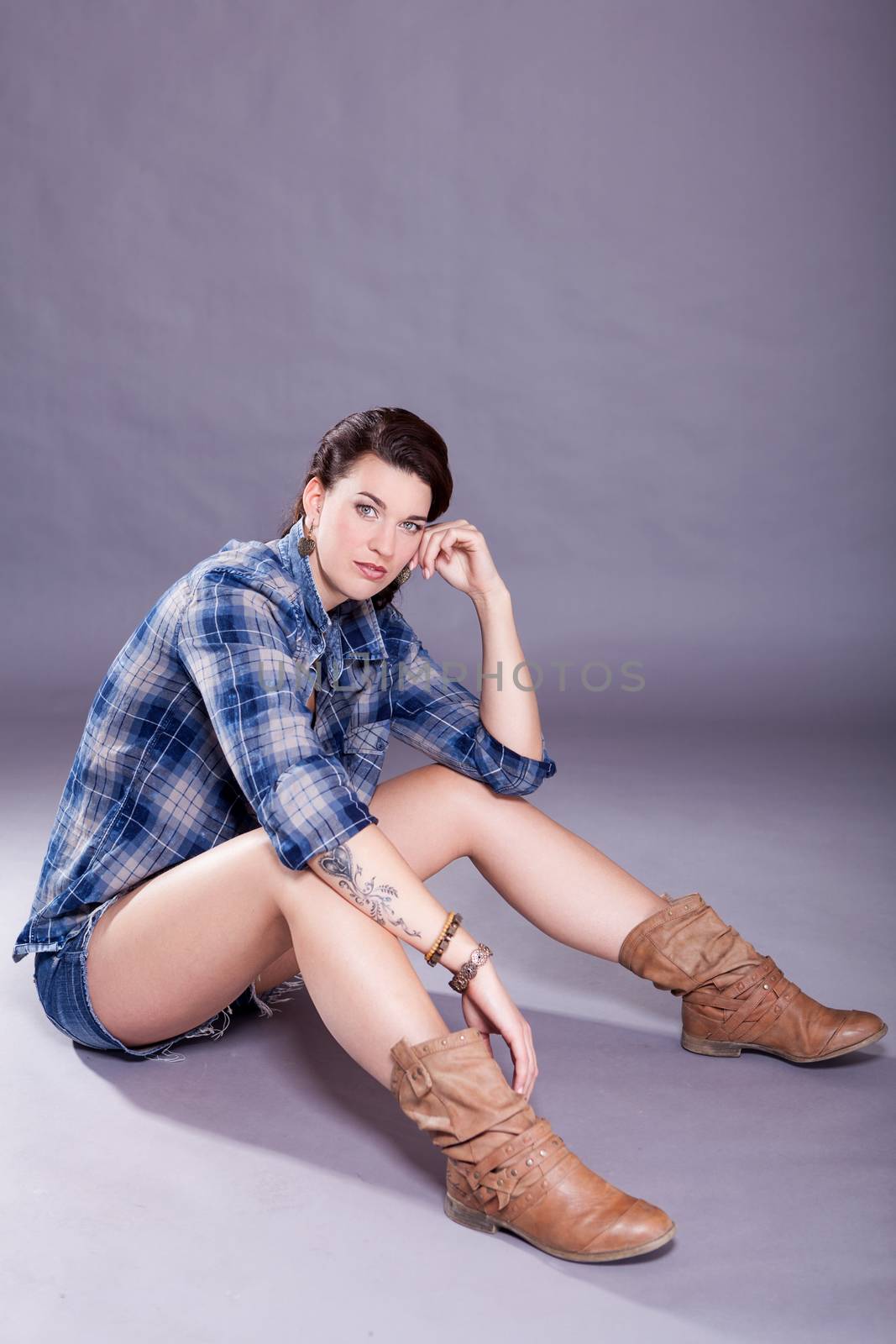 Beautiful single woman in western style boots, jeans shorts and blue flannel shirt with tattooed arm sitting over gray background