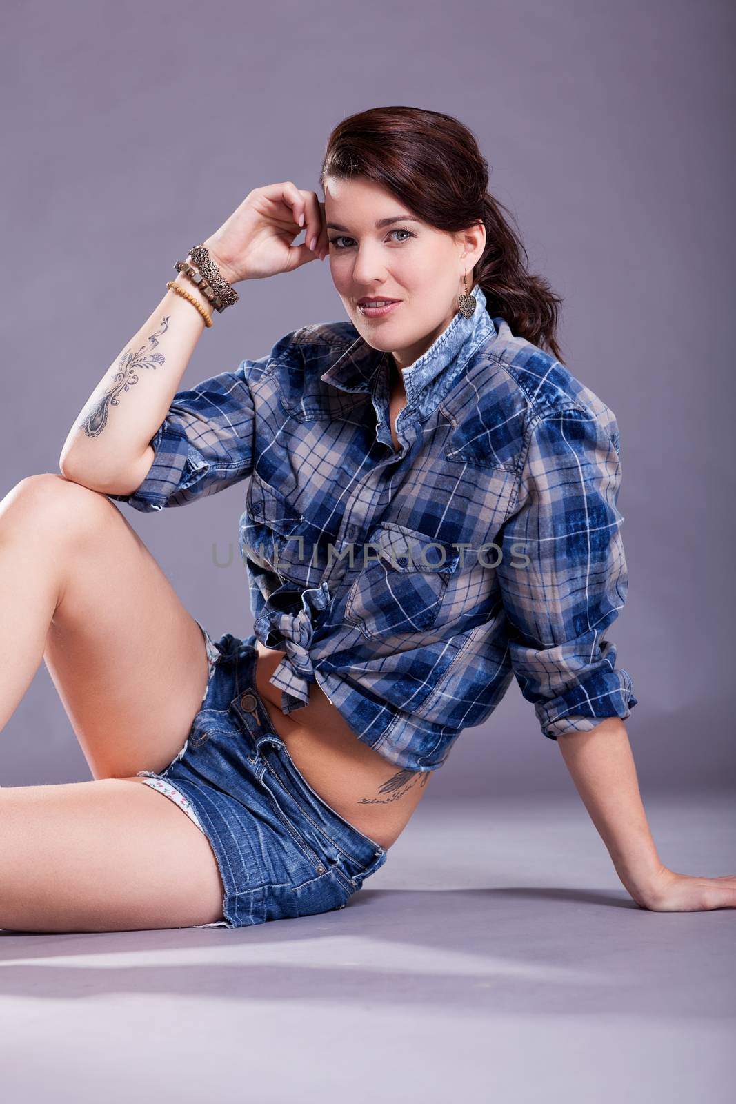 Beautiful single woman in western style boots, jeans shorts and blue flannel shirt with tattooed arm sitting over gray background