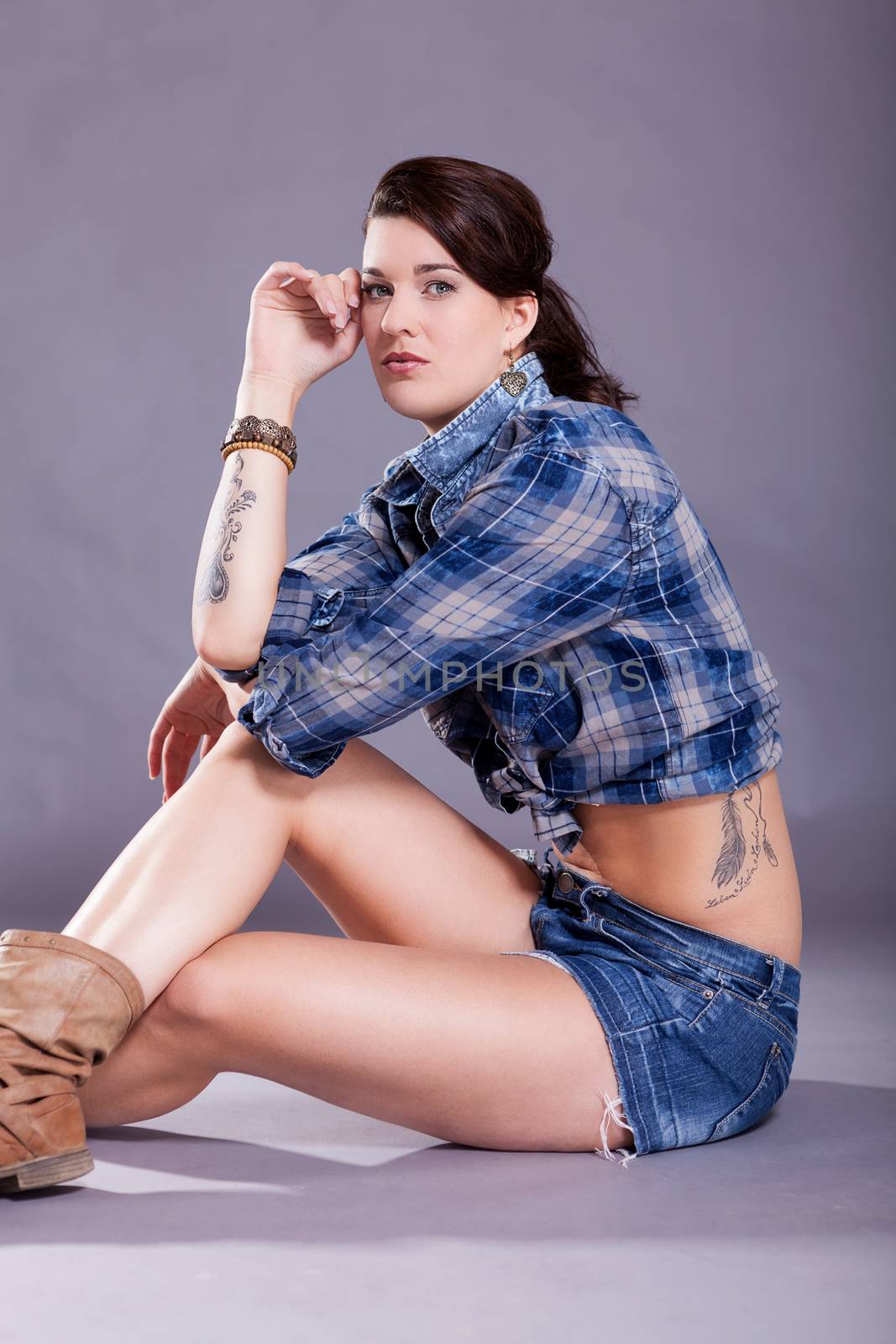 Beautiful single woman in western style boots, jeans shorts and blue flannel shirt with tattooed arm sitting over gray background