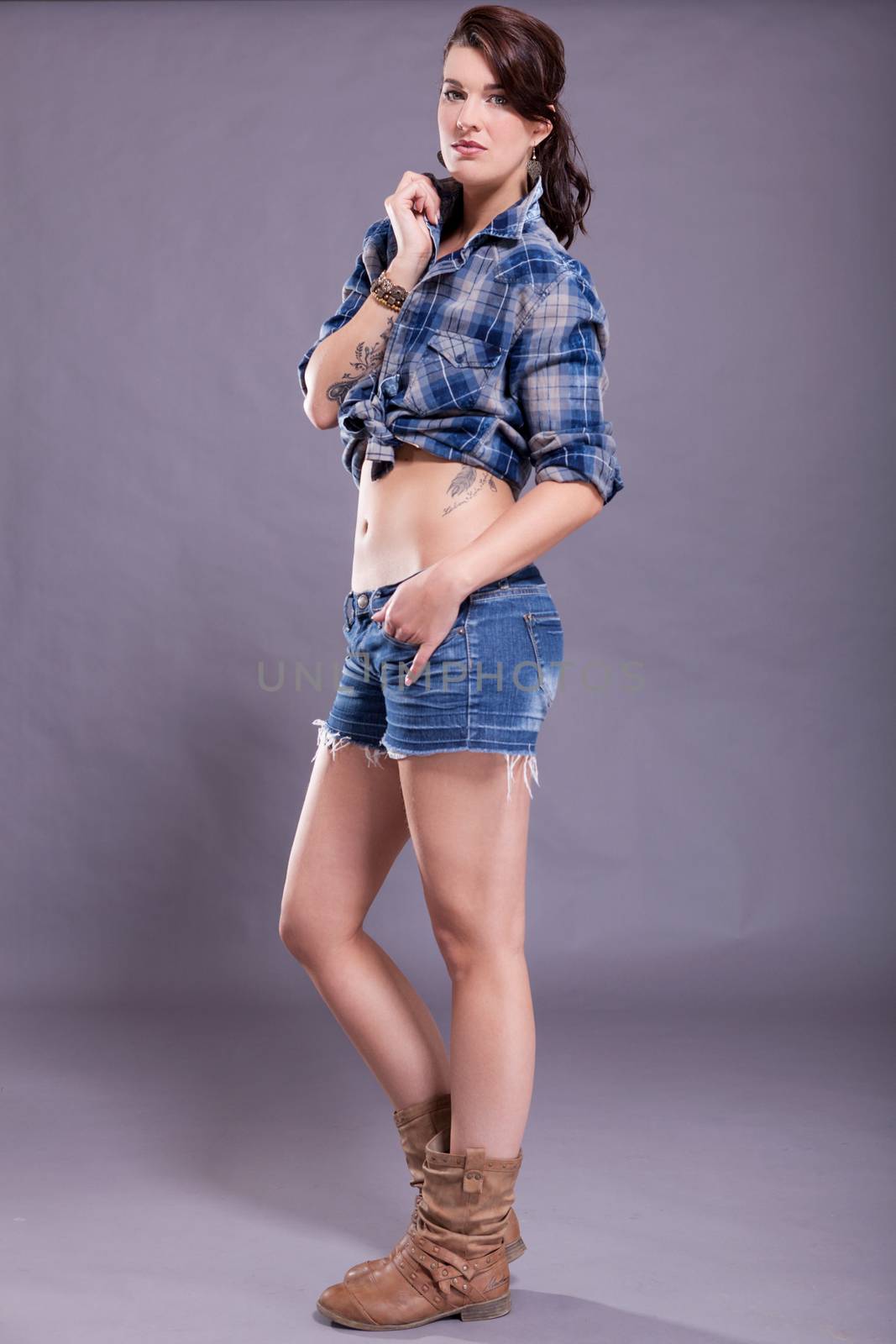 Beautiful single woman in western style boots, jeans shorts and blue flannel shirt with tattooed arm sitting over gray background