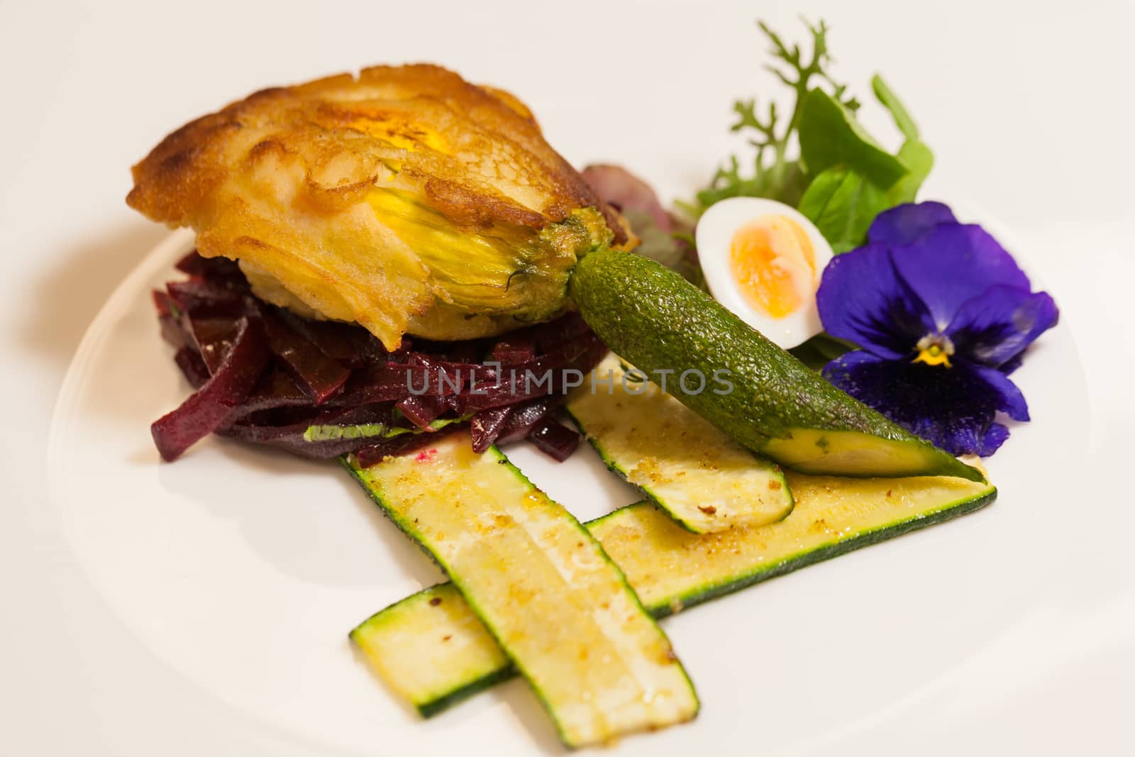Delicious gourmet plate featuring biscuit sandwich with beets, zucchini strips and pansy garnish over white background