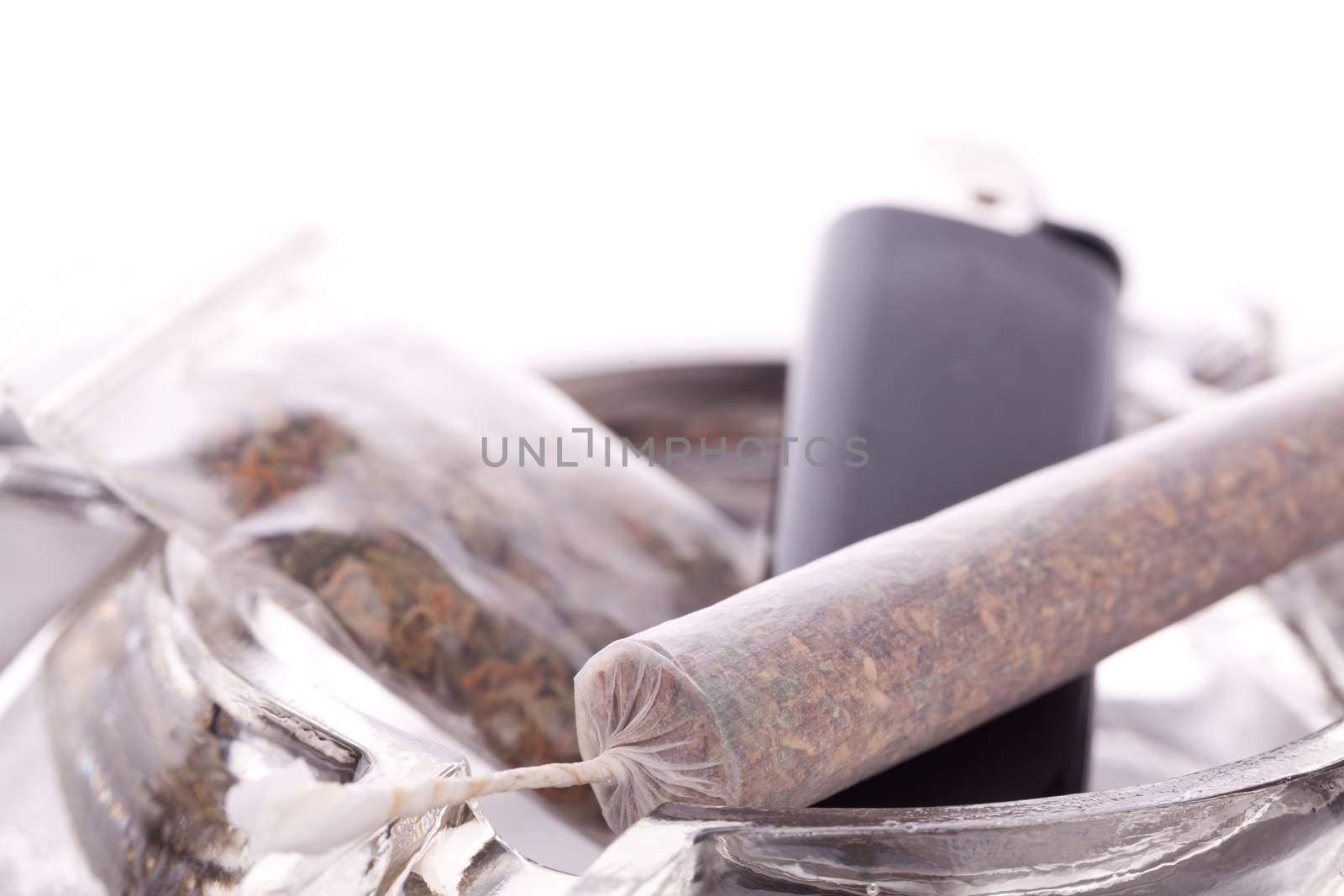 Close up of marijuana joint made with translucent rolling papers, plastic baggy of dried marijuana, black lighter and pipe on white background