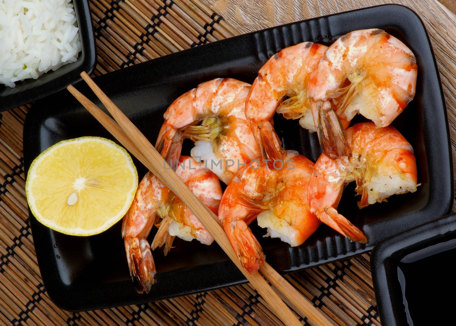 Roasted Shrimps with Lemon and Chopsticks, Soy Sauce and Boiled Rice in Square Bowls on Straw Mat background in Asian Style
