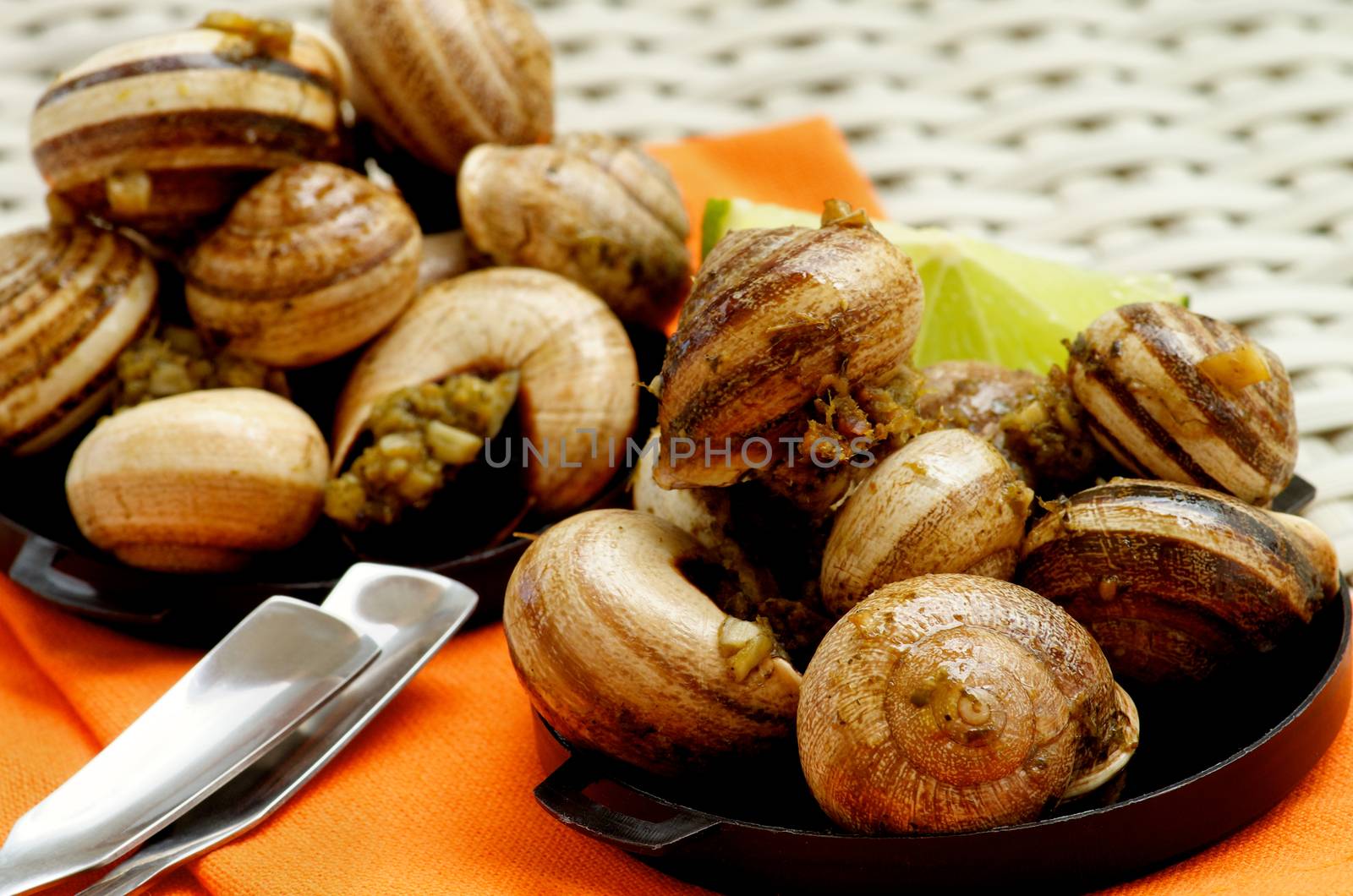 Delicious Escargot with Garlic Butter in Black Bowls with Lime and Snack Spoons on Orange Napkin closeup on Wicker background. Focus on Foreground