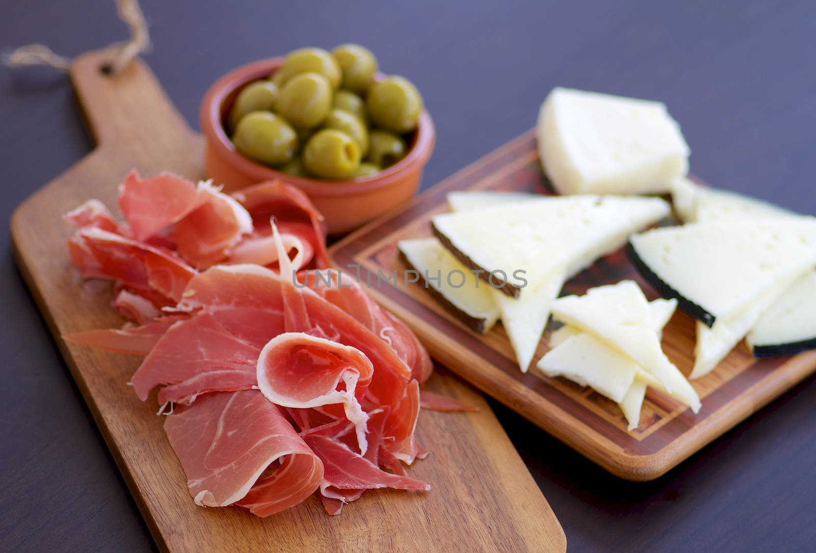 Traditional Spanish Tapas with Sliced Hamon, Goat Cheese and Green Olives on Wooden Cutting Board closeup on Dark Wooden background. Focus on Hamon