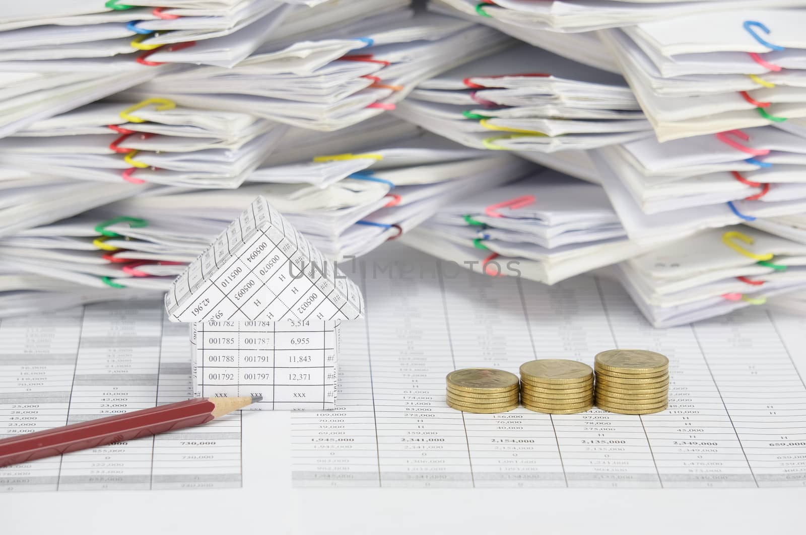 Brown pencil and house with step pile of gold coins on finance account have blur  overload of paperwork with colorful paperclip as background.