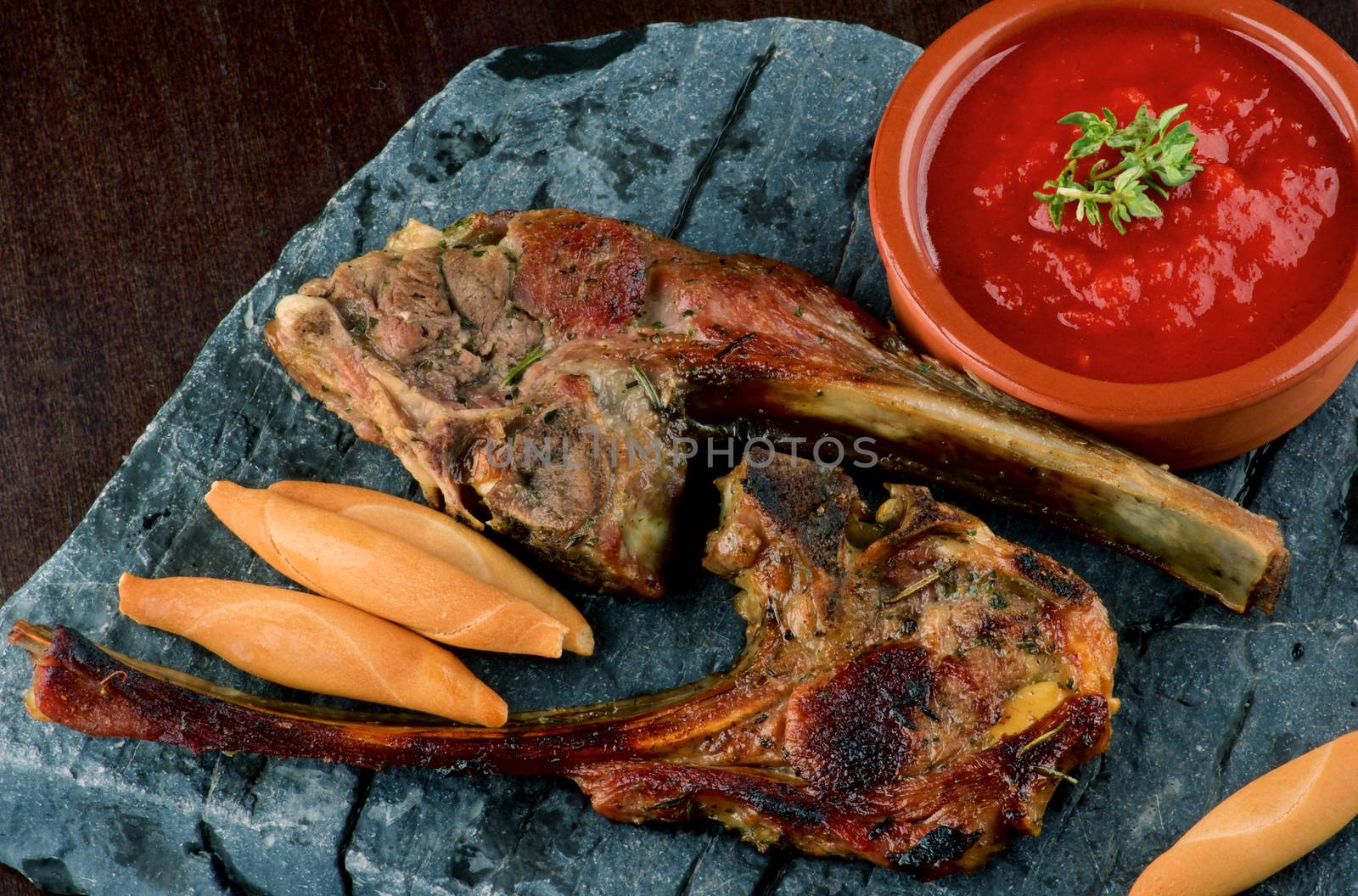 Delicious Roasted Lamb Ribs with Bread Sticks and Tomato Sauce closeup on Stone Plate