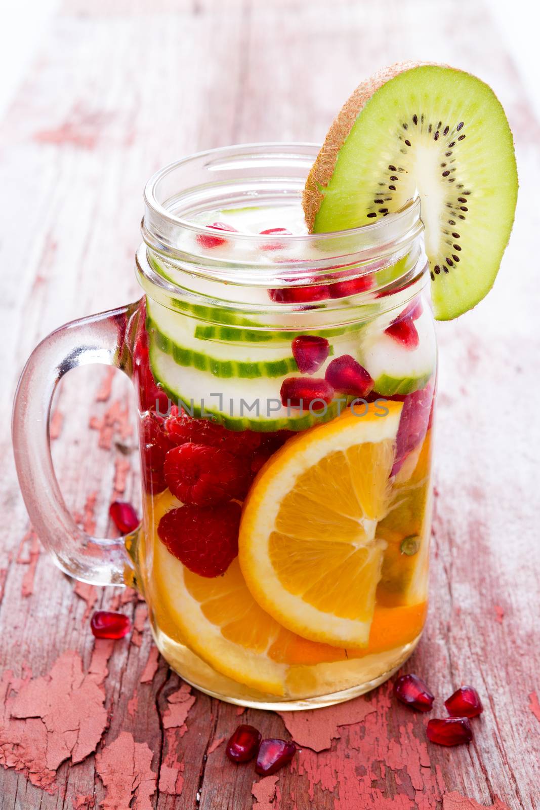 Fresh fruit and water filled mason jar with sliced oranges, cucumber, raspberries, pomegranate and kiwifruit for a delicious refreshing healthy drink on a rustic wooden table with peeling paint