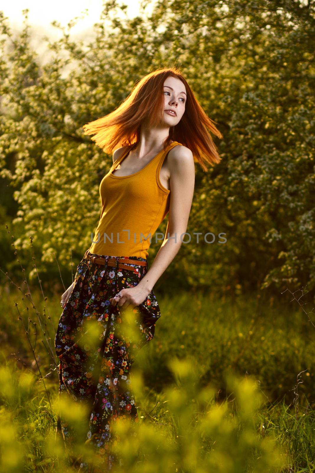 Beautiful redhead girl casually walking under the rays of the sunset in summer forest